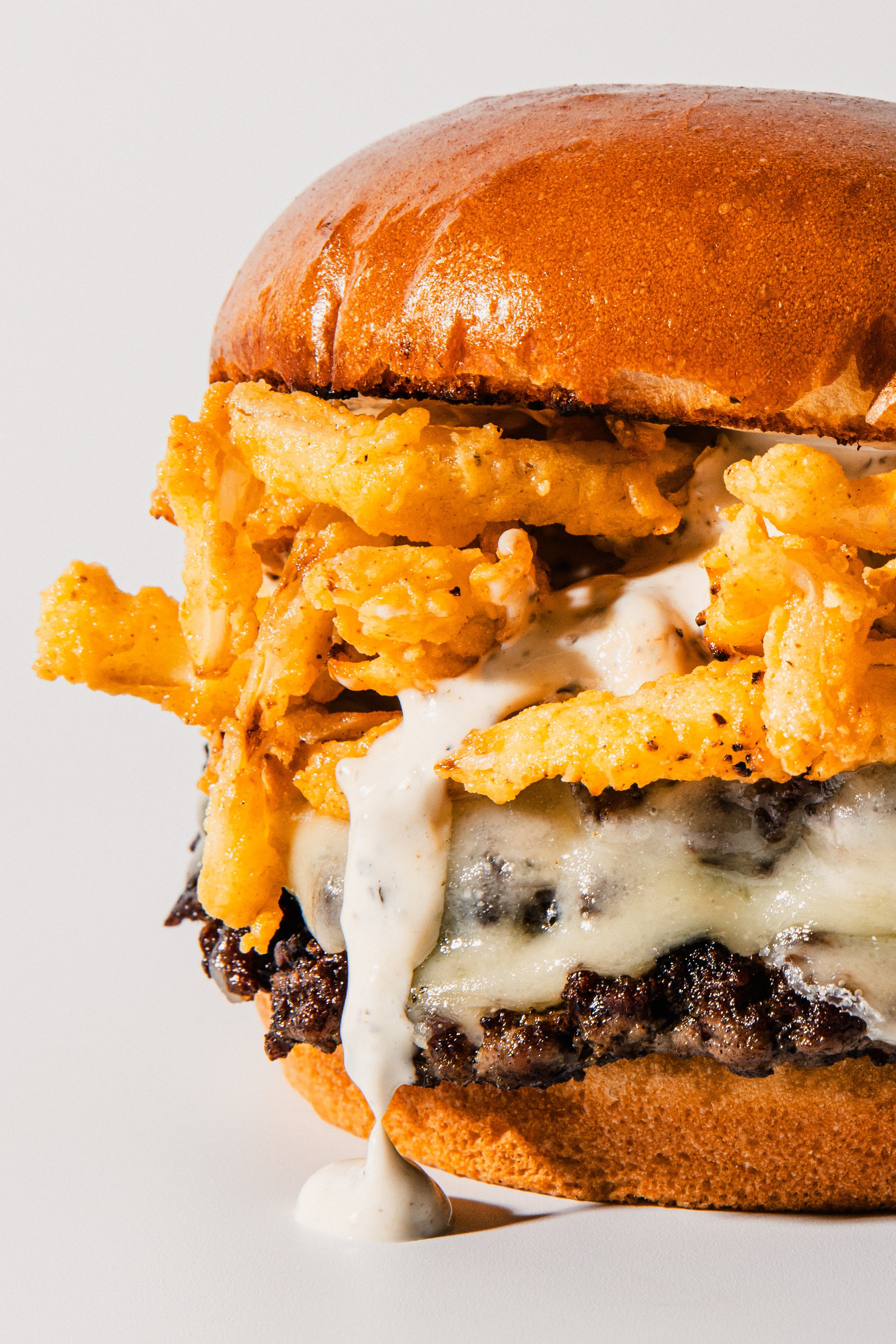 A close-up of a burger with a sesame seed bun, topped with crispy fried onion strings, melted cheese, and a beef patty with a drizzle of white sauce.