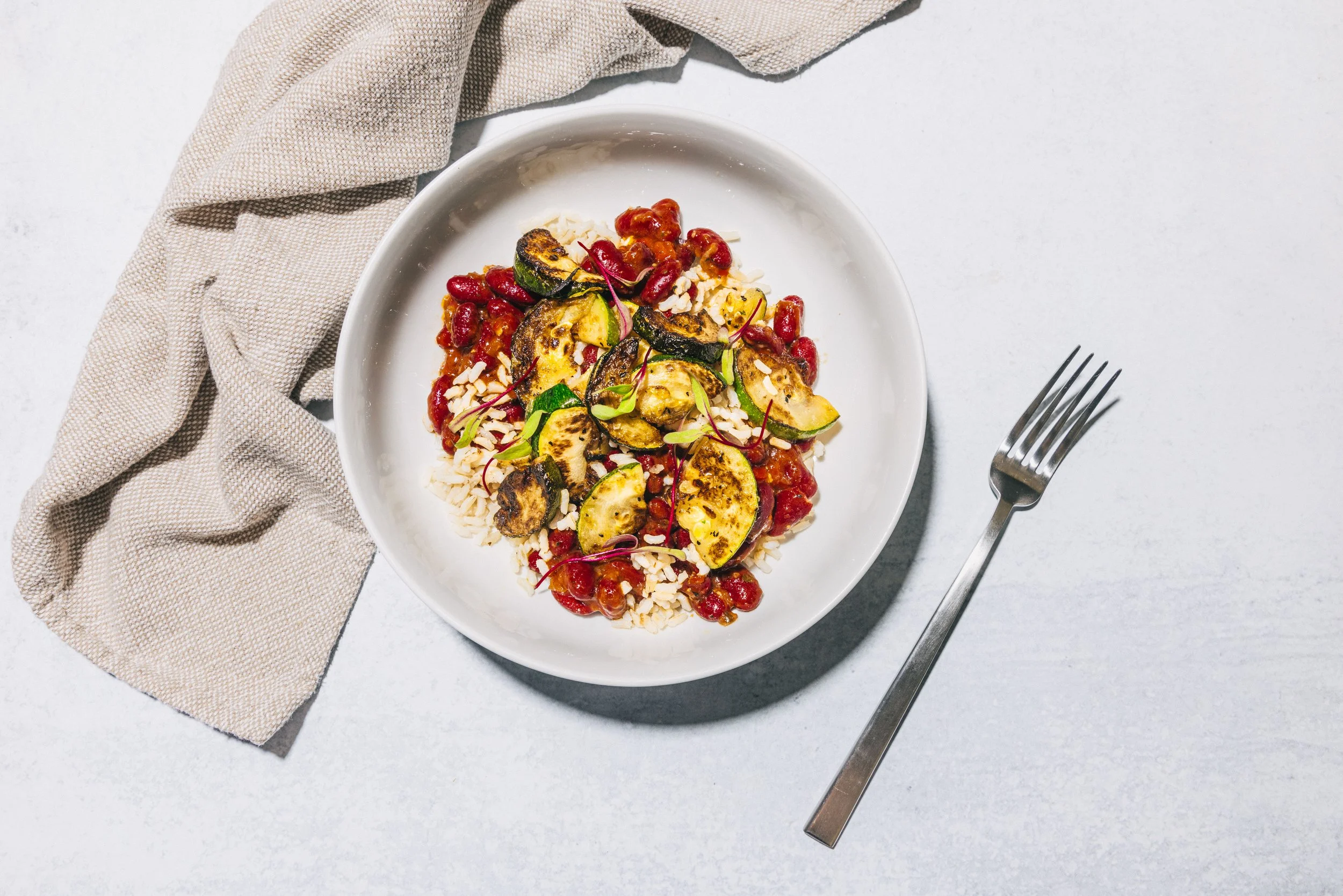 A white bowl filled with roasted squash, pomegranate seeds, shredded cheese, and microgreens, set on a white surface with a beige cloth and a fork nearby.