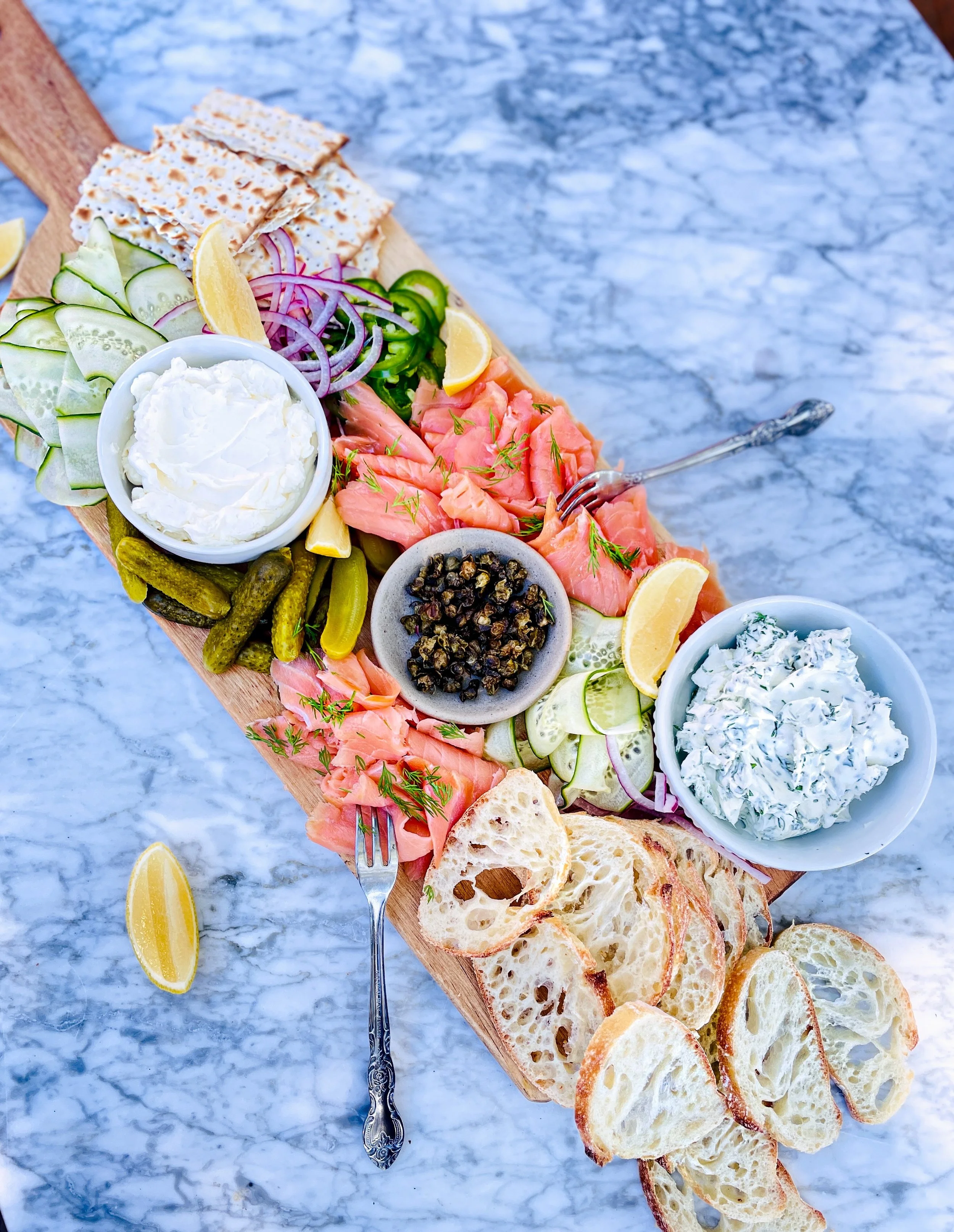 A wooden serving board with slices of toasted bread, sliced cucumbers, red onions, lemon wedges, smoked salmon, and bowls of cream cheese and capers on a marble table.