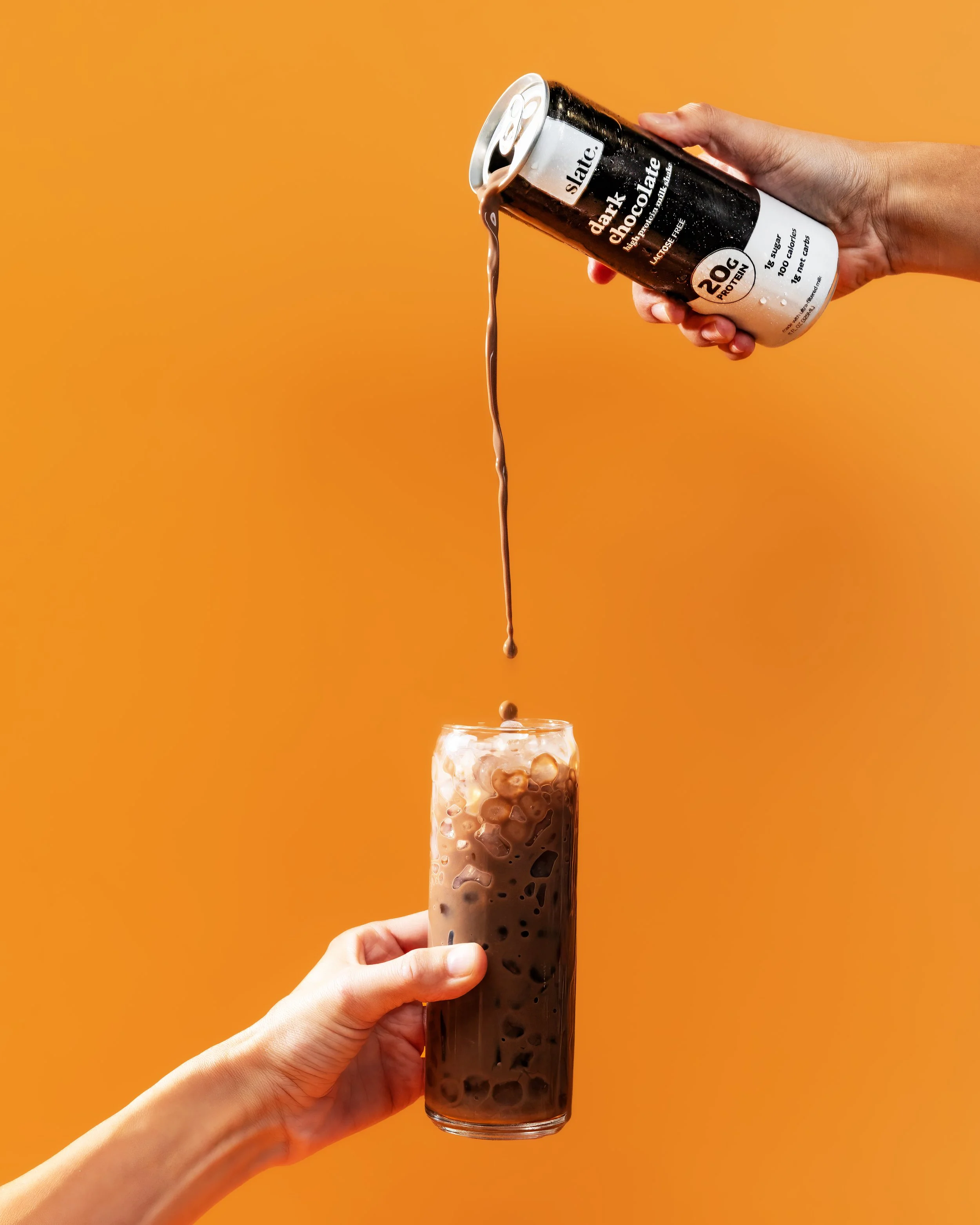 Hand holding a tall glass of iced coffee with ice cubes, over an orange background, with another hand pouring chocolate soda from a black can into the glass.
