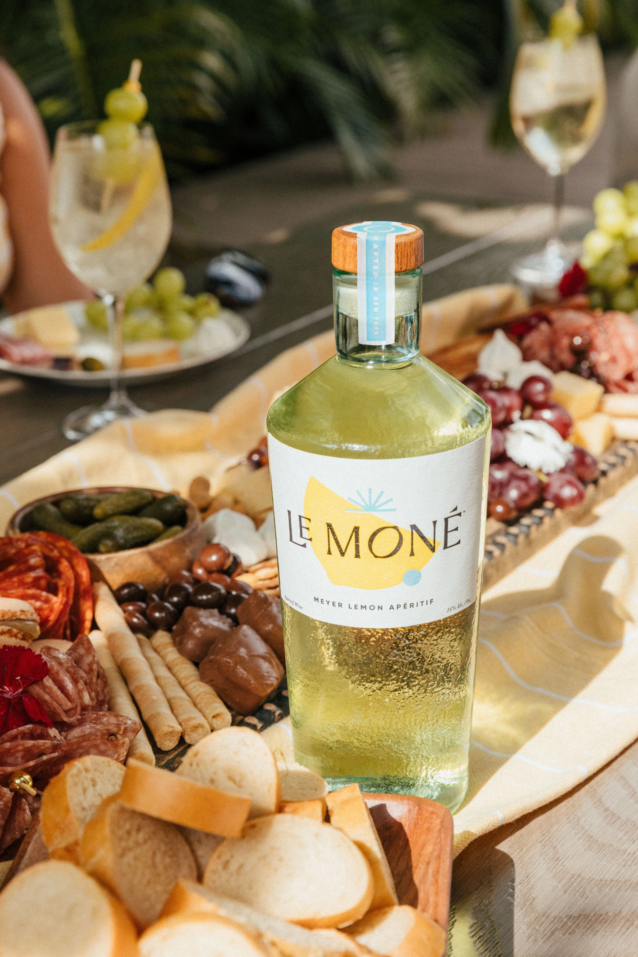 A bottle of Le Moné Meyer Lemon Aperitif on a table with a cheese and charcuterie platter in the background, including grapes, sliced meat, cheese, and crackers, with glasses of wine nearby.