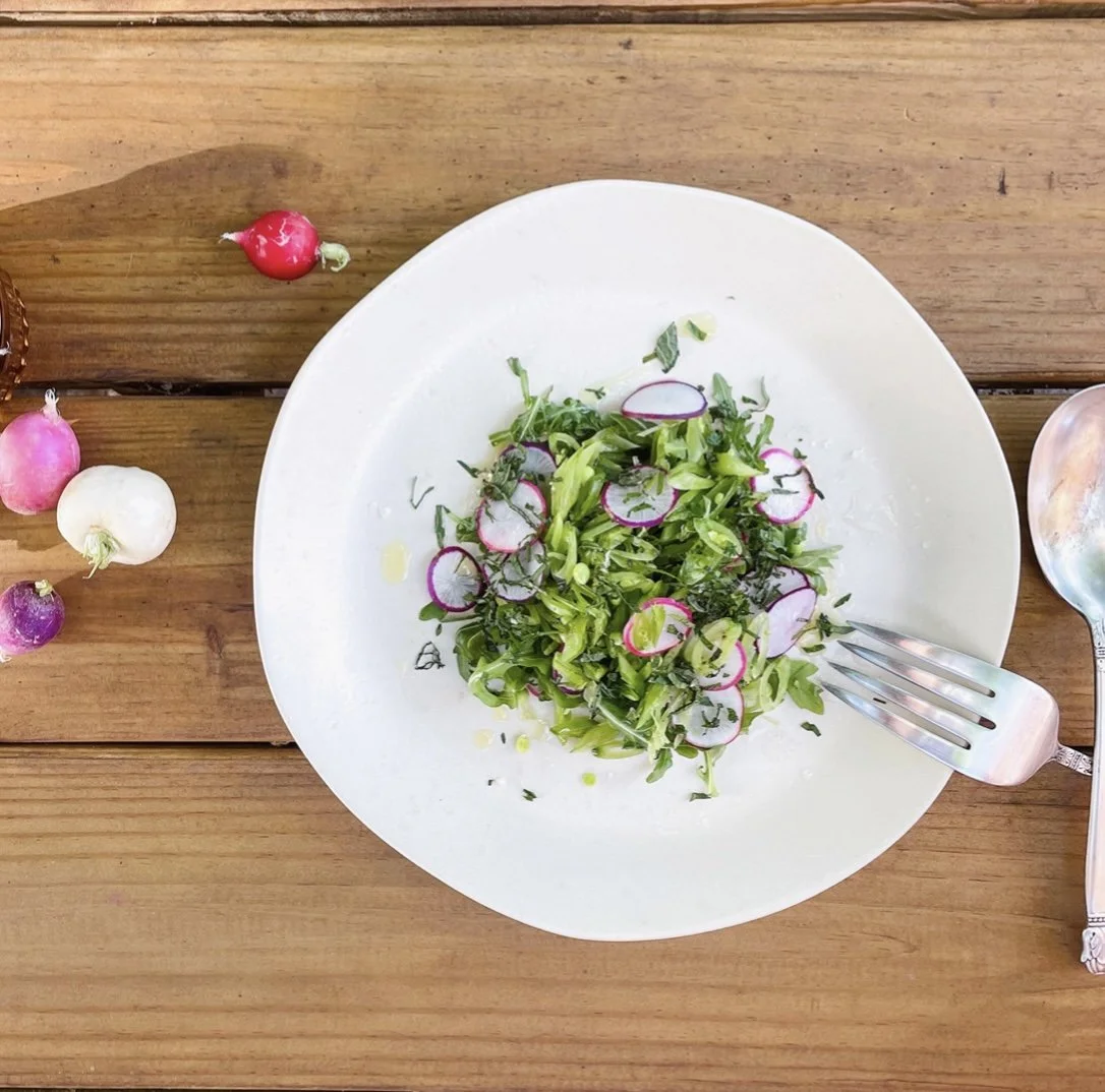 A plate of mixed fresh greens topped with sliced radishes on a wooden table, with whole radishes scattered nearby and a spoon partially visible.