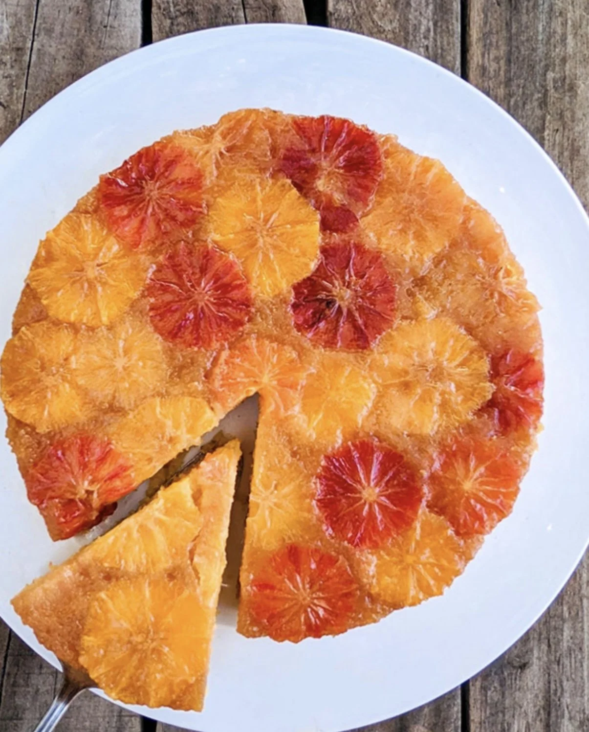 Round cake topped with sliced blood oranges and regular oranges, with one slice cut out, on a white plate on a wooden surface.