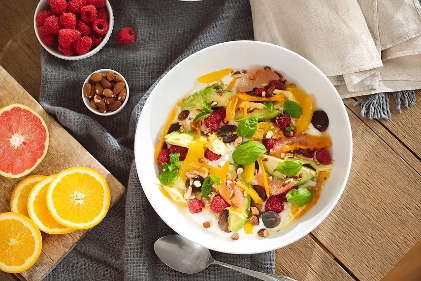 A bowl of salad with mixed greens, raspberries, black olives, shredded cheese, and sunflower seeds on a wooden table. Nearby are sliced oranges, a halved grapefruit, a bowl of raspberries, and another bowl with almonds.