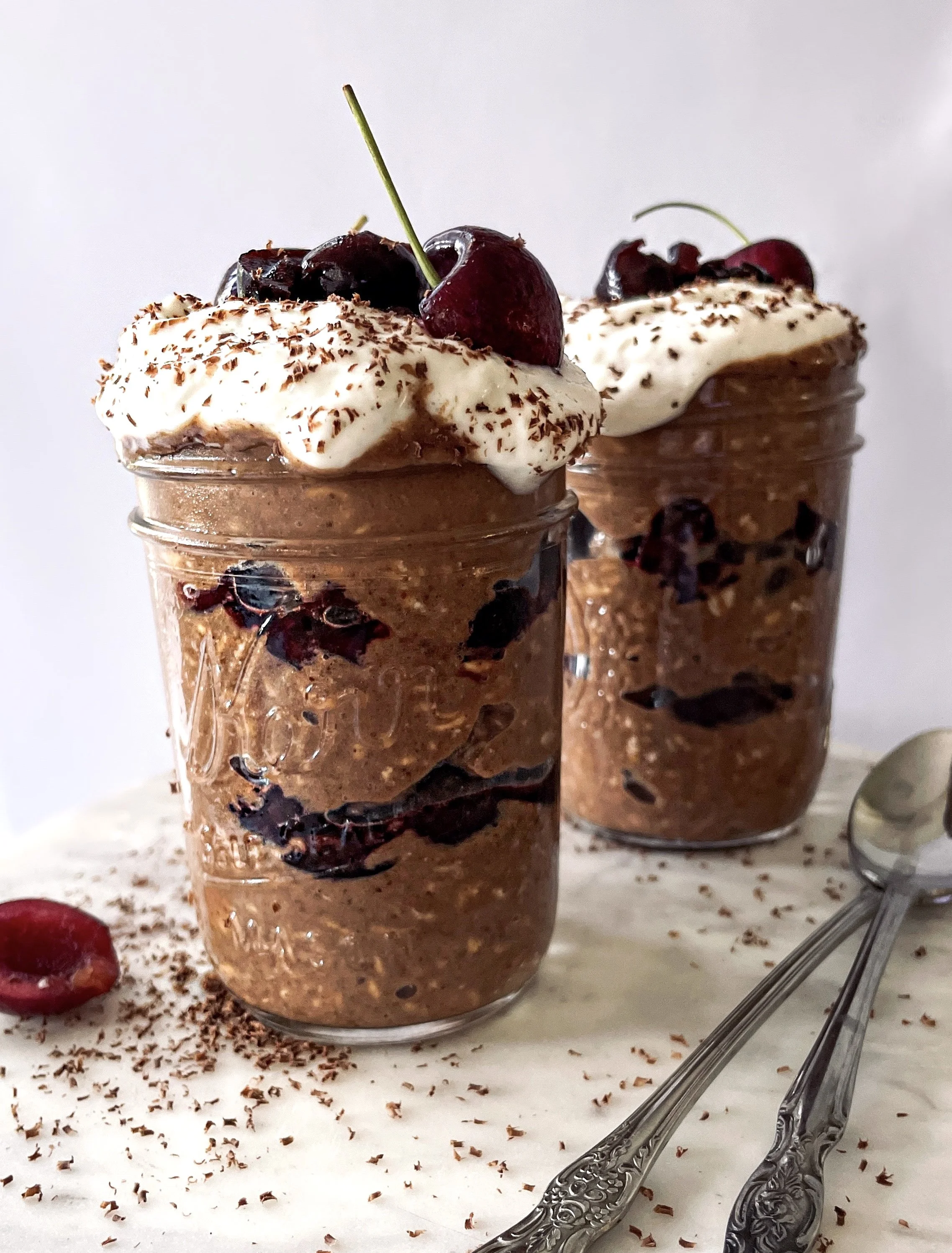 Two glass jars filled with layered chocolate mousse topped with whipped cream, cherries, and chocolate shavings, with spoons beside them and chocolate shavings scattered on the surface.