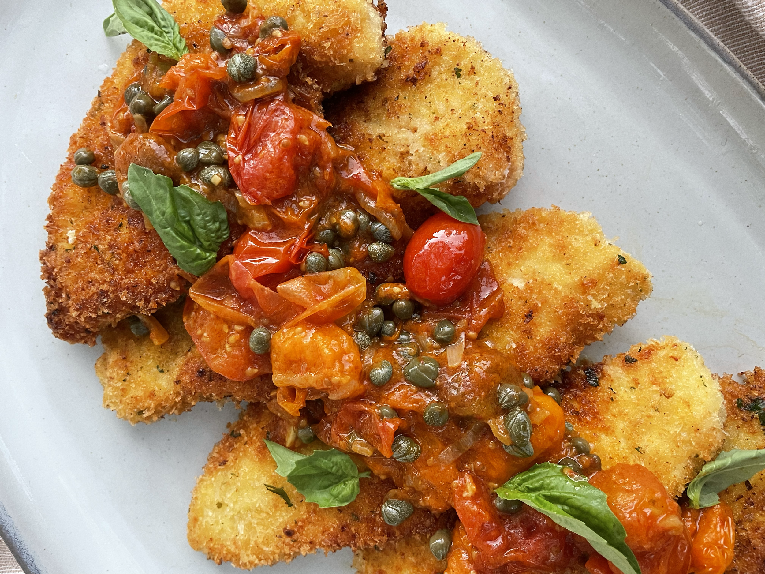 Close-up of breaded fish fillets topped with tomato-based sauce, cherry tomatoes, capers, and fresh basil leaves on a white plate.