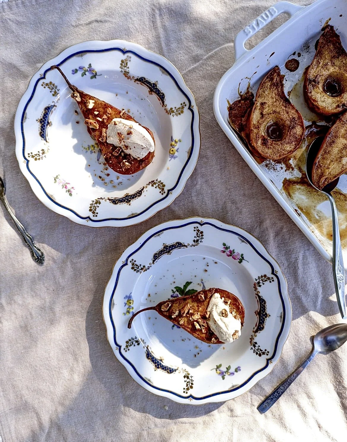 Two plates with poached pears topped with whipped cream and chopped nuts, and a baking dish with baked pears, on a table with a beige tablecloth.