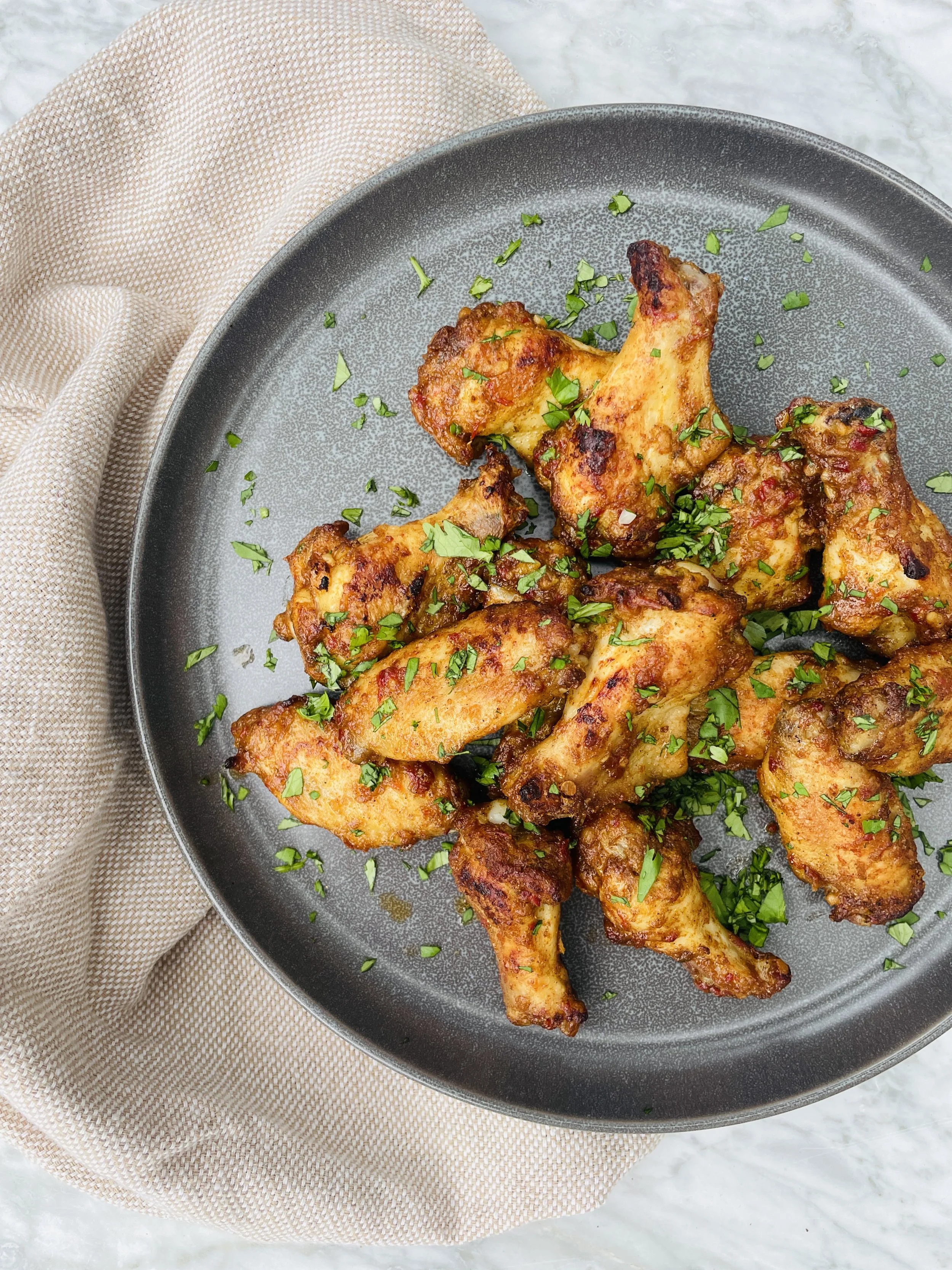 Plate of cooked chicken drumsticks garnished with chopped green herbs, on a textured gray plate, with a beige cloth nearby.