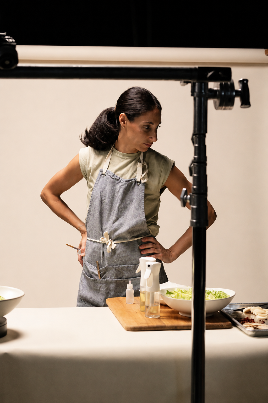 A woman chef in a gray apron and beige shirt preparing food in a kitchen studio with a white background.