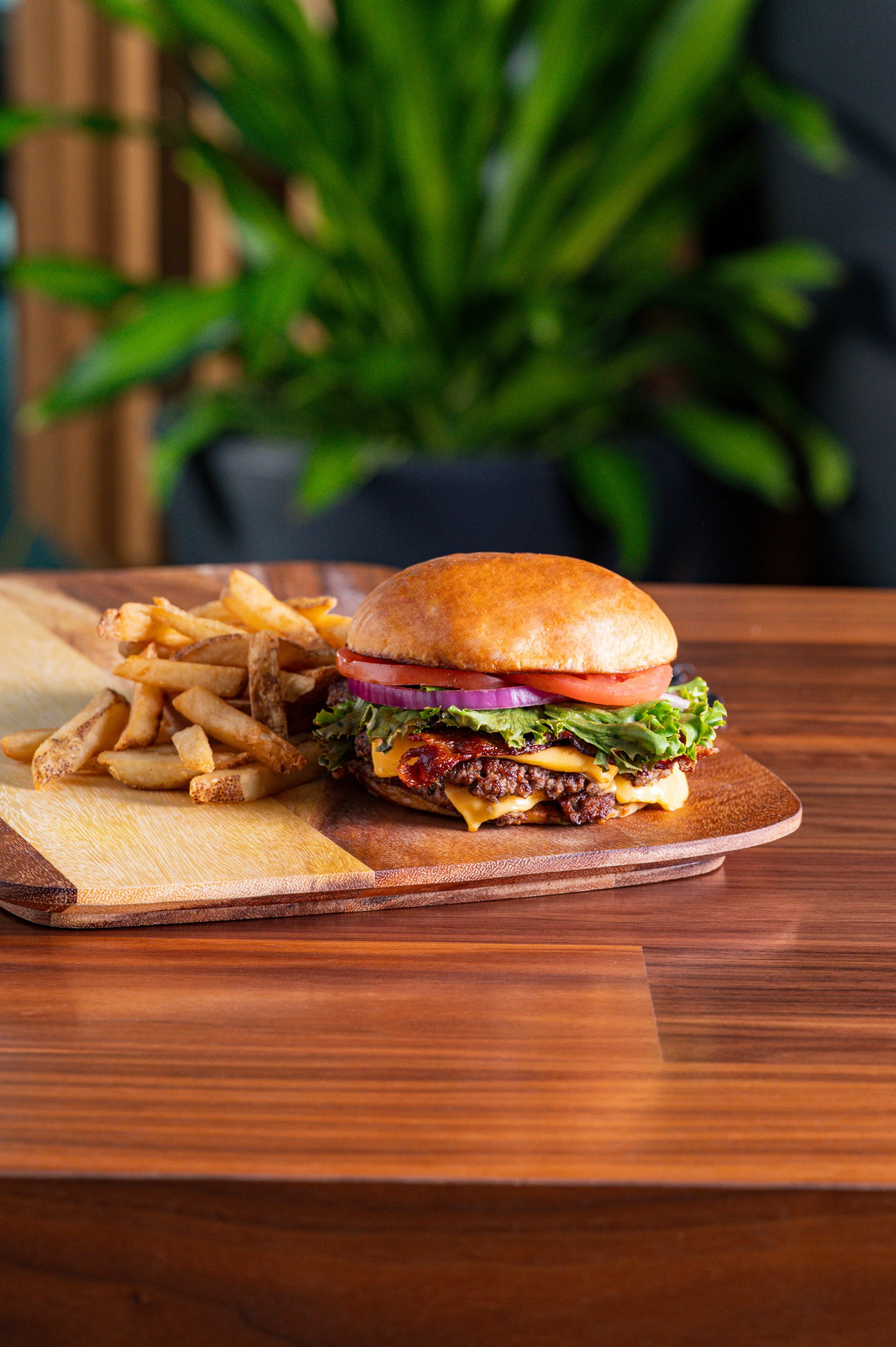 Close-up of a cheeseburger with lettuce, tomato, onion, bacon, and melted cheese on a bun, served with French fries on a wooden platter, with a potted plant in the background.