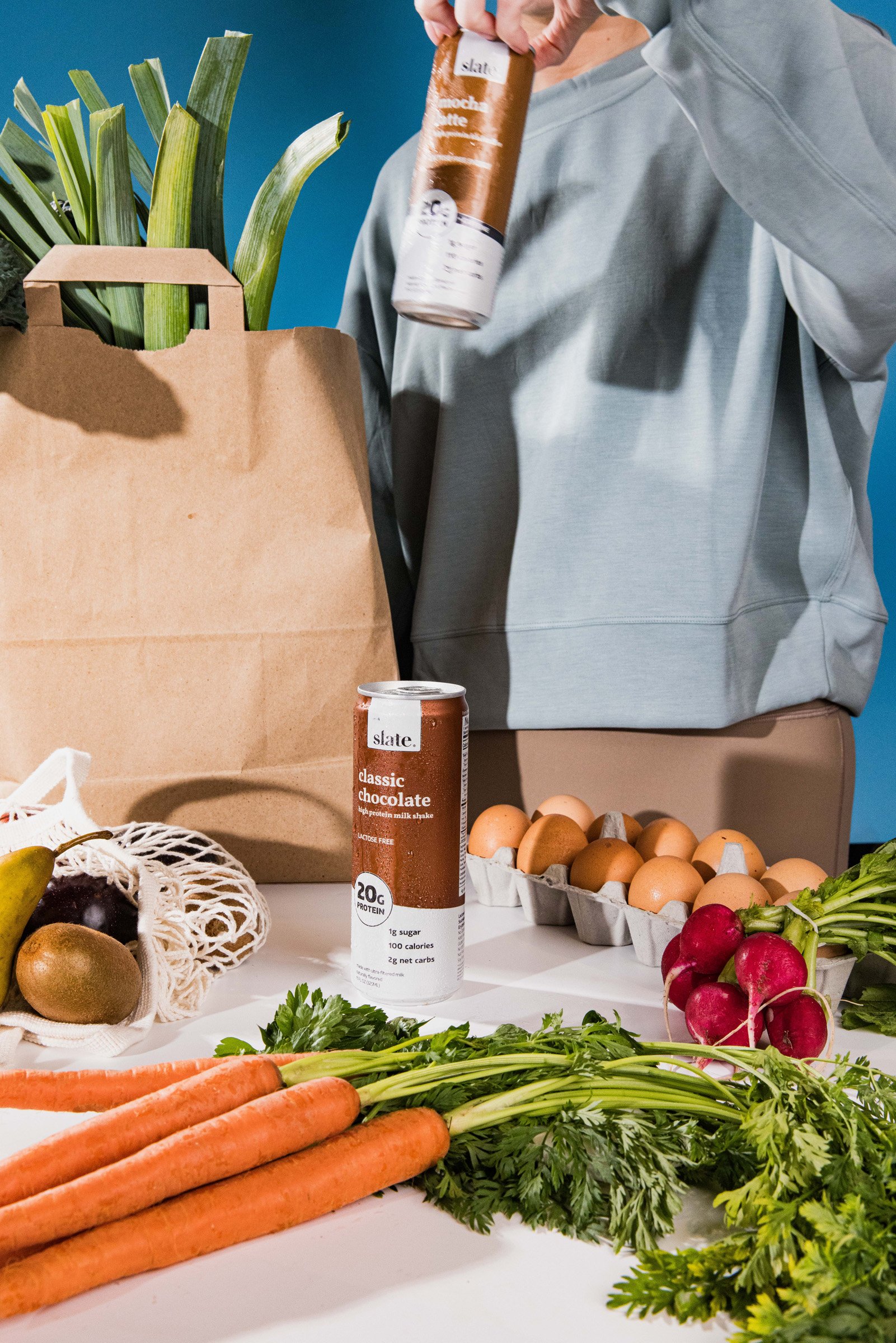 A person in a gray hoodie standing behind a kitchen counter with fresh vegetables, eggs, and a can of chocolate protein shake. The counter also has a brown paper grocery bag with green leeks inside and various fruits like carrots, radishes, and grape