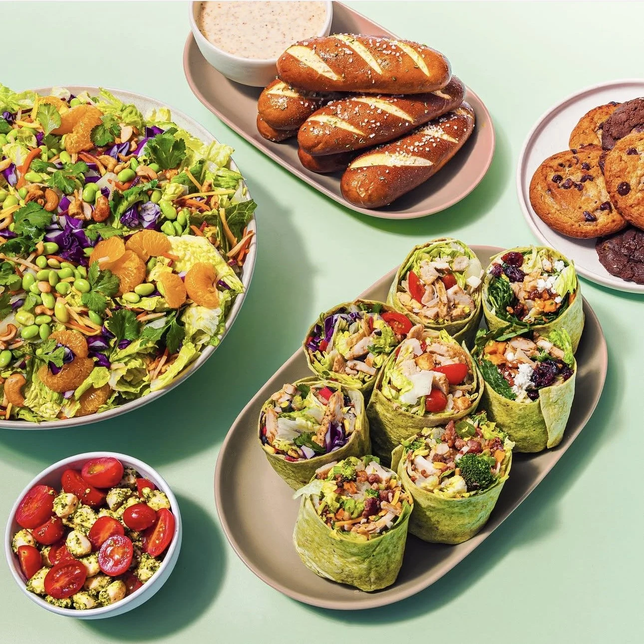 A variety of colorful dishes including a large salad with greens and mandarin oranges, a plate of grilled sausages with mustard, a bowl of chocolate chip cookies, a plate of veggie wraps with various vegetables, a small bowl of cherry tomatoes and fe
