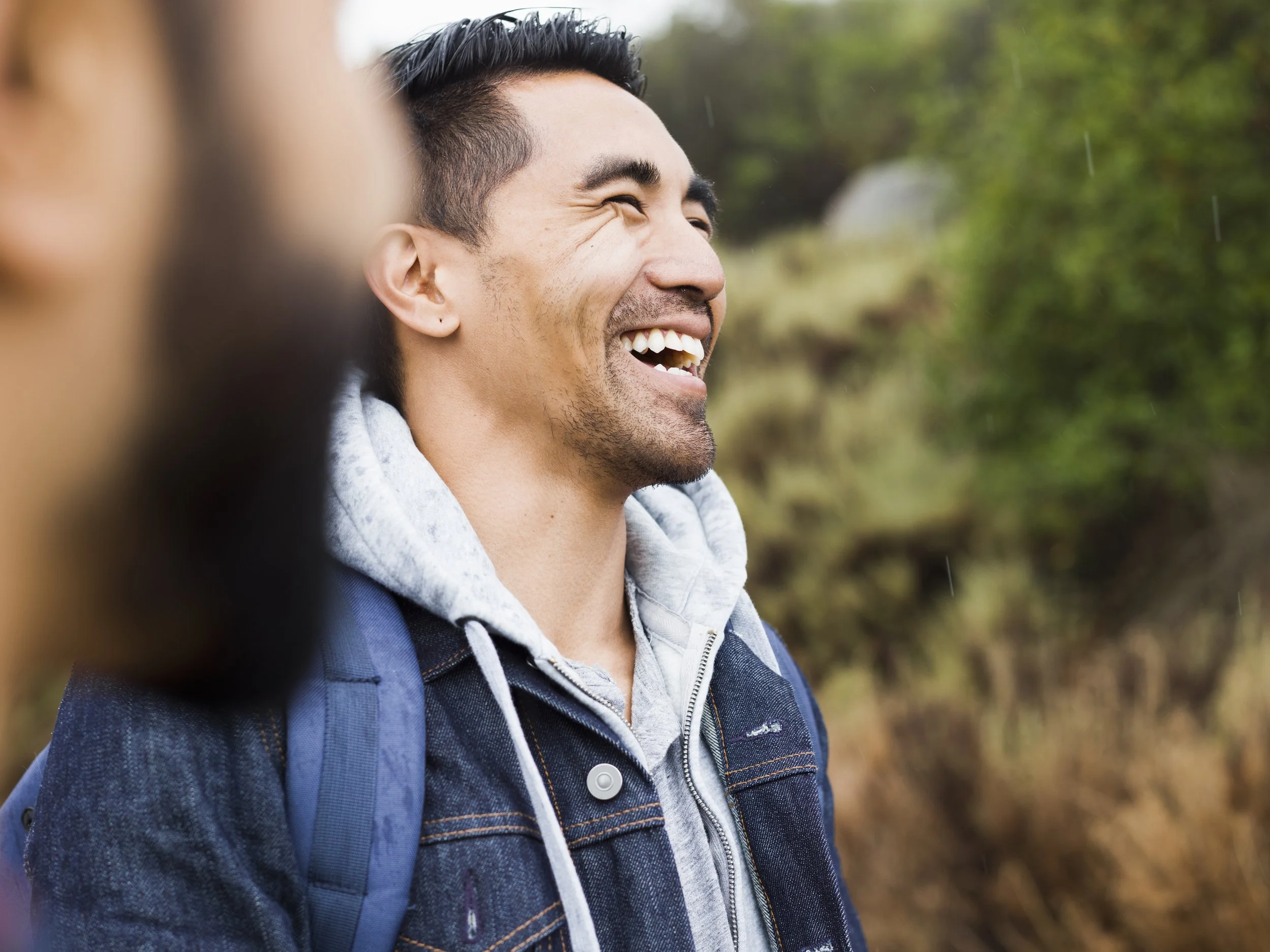 A man outdoors smiling and laughing, wearing a denim jacket and gray hoodie, with a backpack on a trail surrounded by trees.