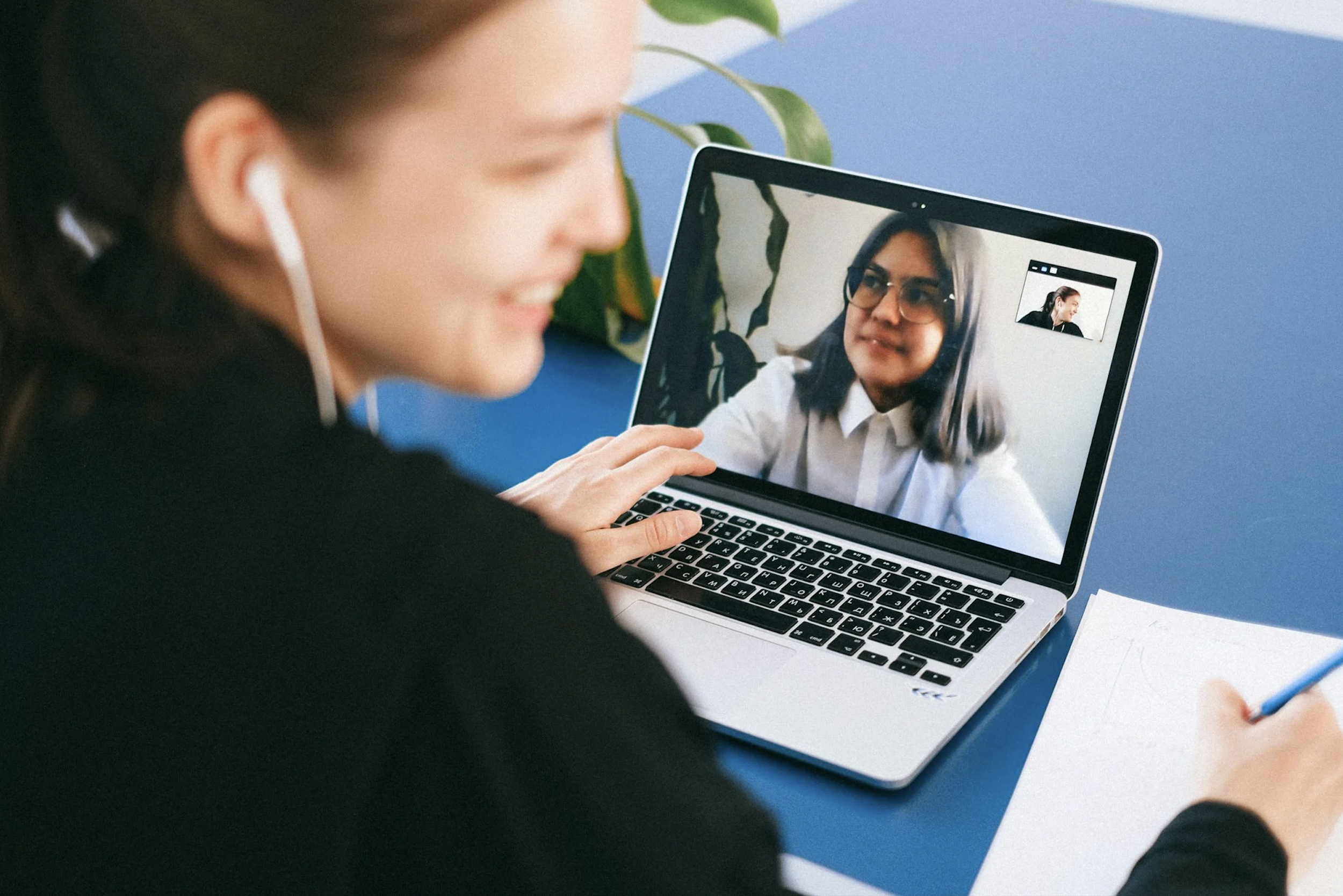 Person with earbuds on a video call on a computer and taking notes.