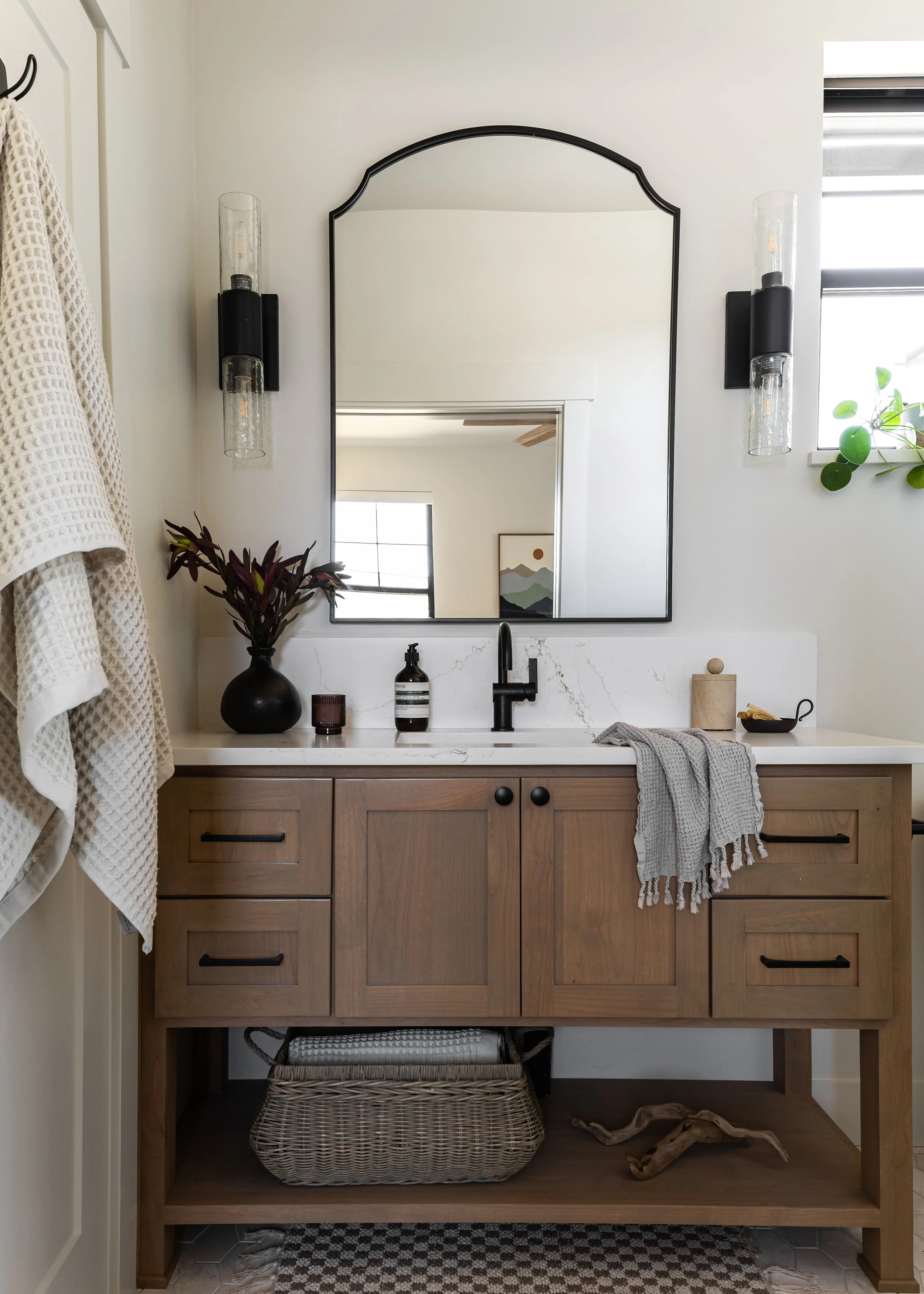 A bathroom vanity with a white marble countertop and a wooden cabinet below. A black vase with dark purple leaves, soap dispenser, candle, and other small items on the countertop. A large mirror above the vanity and modern wall sconces on each side. A small window with a green plant on the right and a patterned rug underneath.