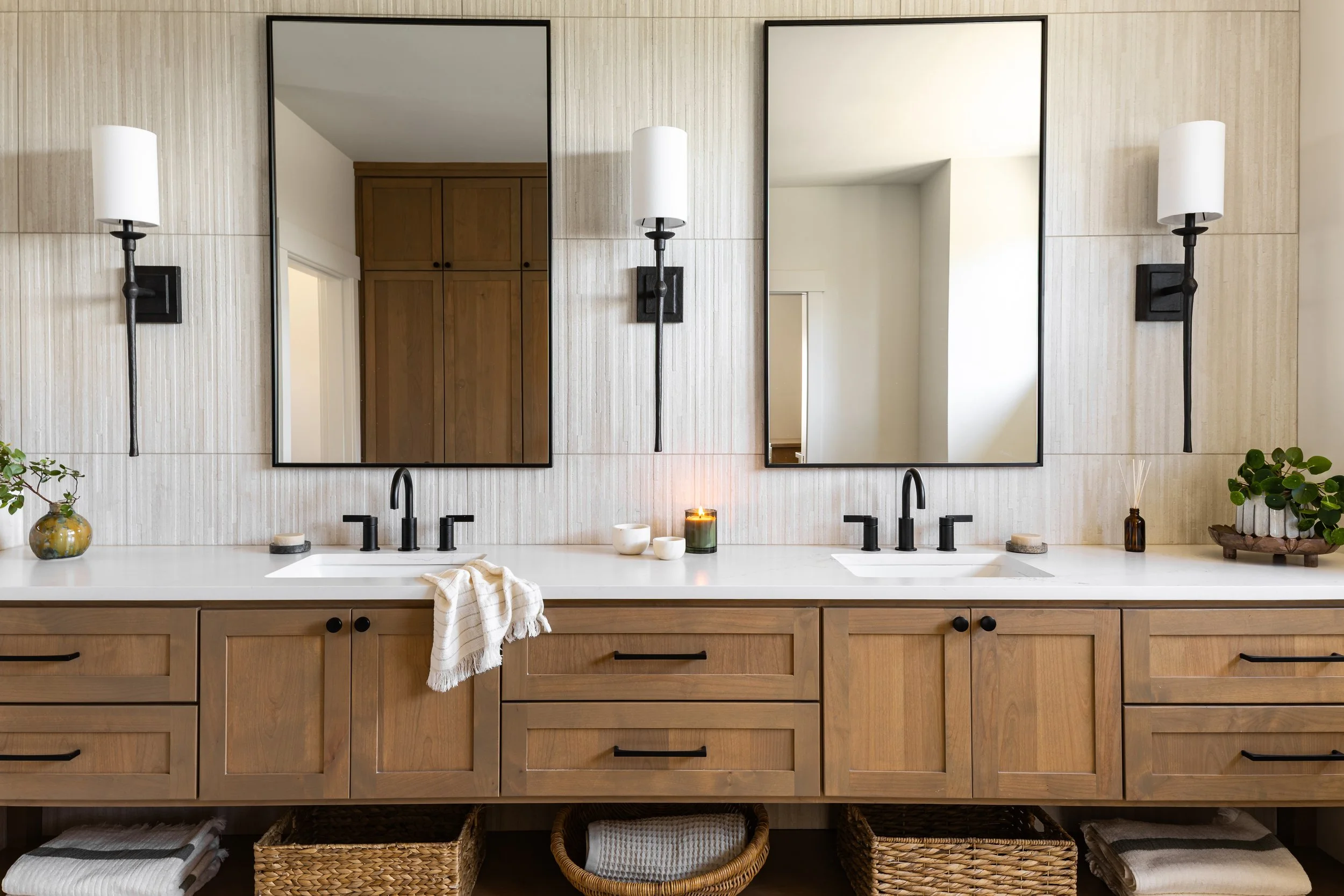 Bathroom vanity with double black faucets, two large mirrors, and wall-mounted light fixtures. Decor includes small vases with greenery and candles, with woven baskets and towels stored underneath.