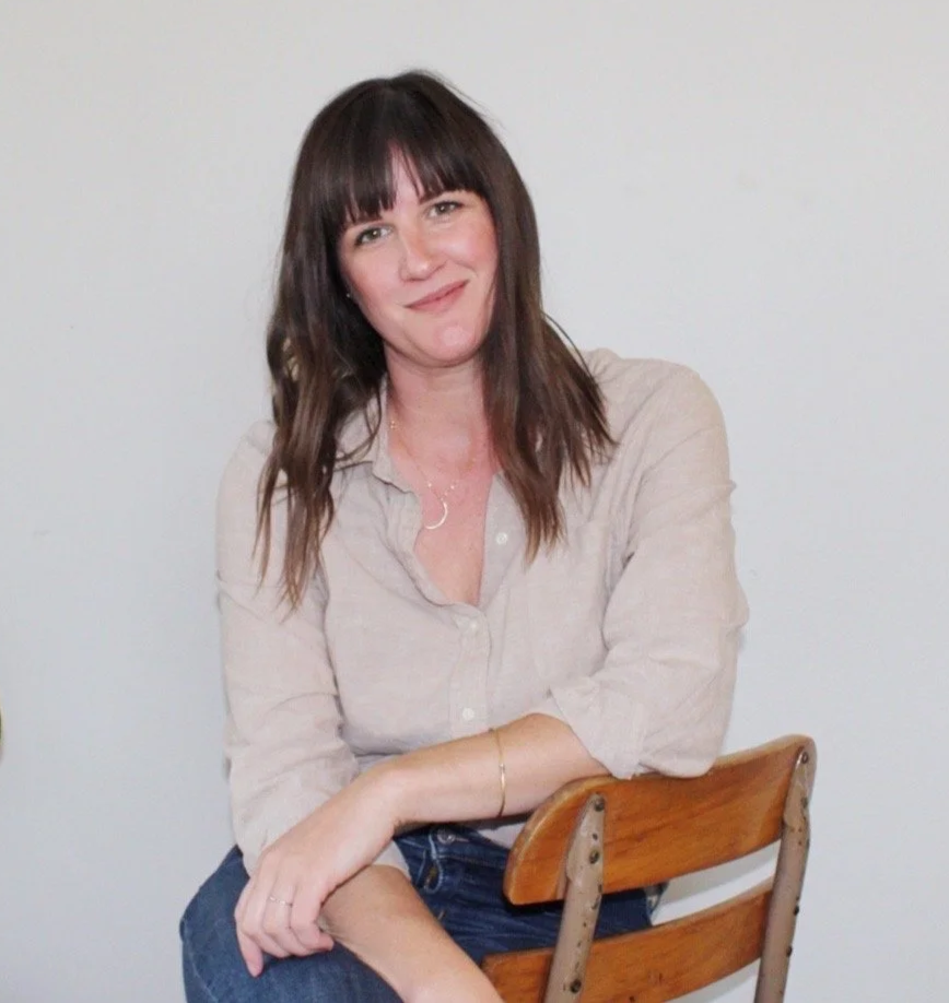 A woman with shoulder-length dark brown hair and bangs, smiling, wearing a light beige button-up shirt with rolled-up sleeves, sitting on a wooden chair with a curved backrest, against a plain white wall.
