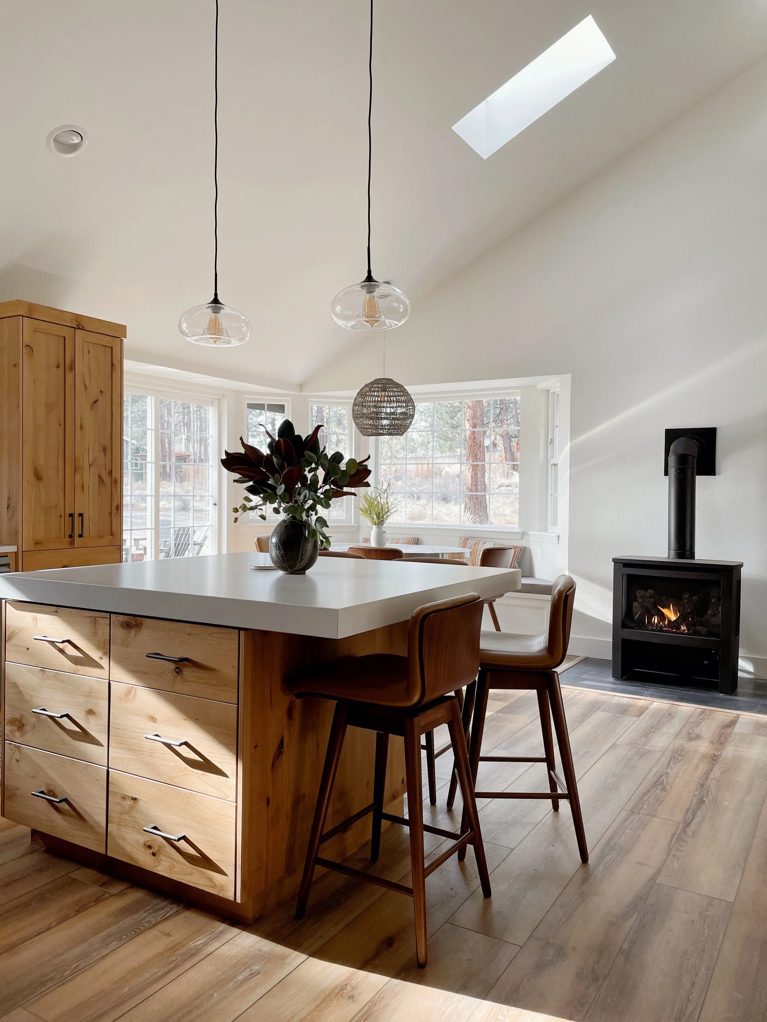 Modern kitchen with a wooden island, a white countertop, wooden chairs, large windows, and a fireplace with a chimney, during daylight.