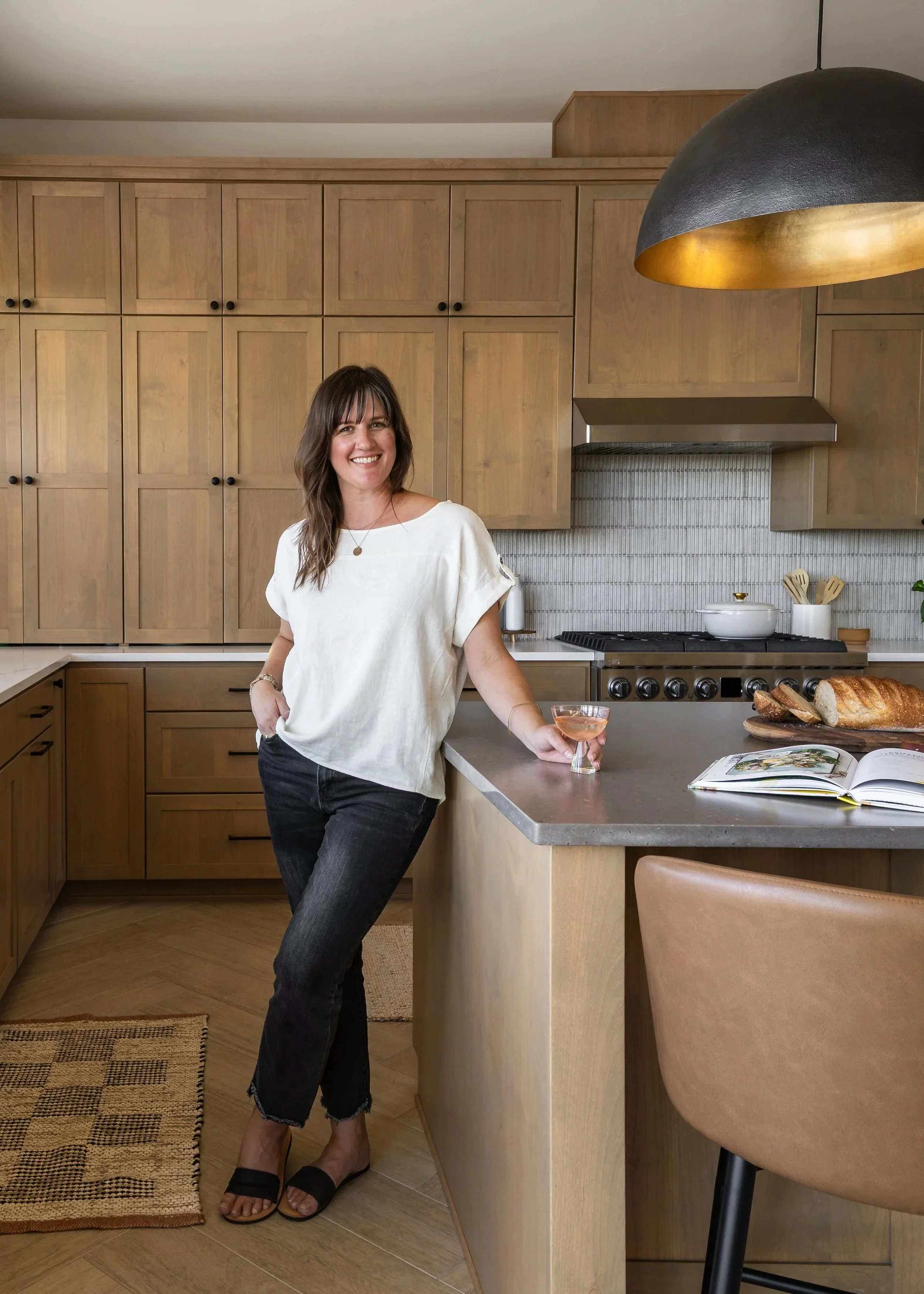 A woman in a white shirt and black pants standing in a kitchen, smiling, holding a glass of rosé wine, with bread and an open book on the counter.