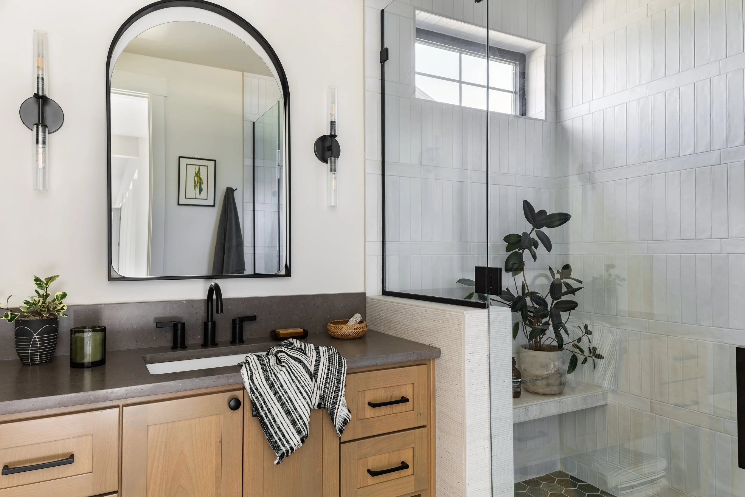 Modern bathroom with wooden vanity, black fixtures, large mirror, glass shower with a window, indoor plants, and minimal decor.