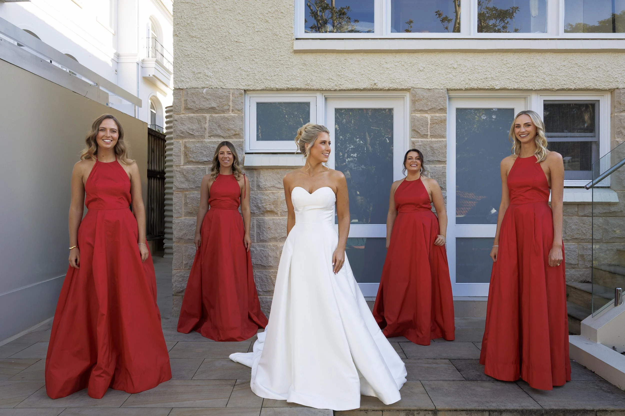 A bride in a white wedding dress with her bridesmaids in red dresses standing outdoors in front of a beige stone building.