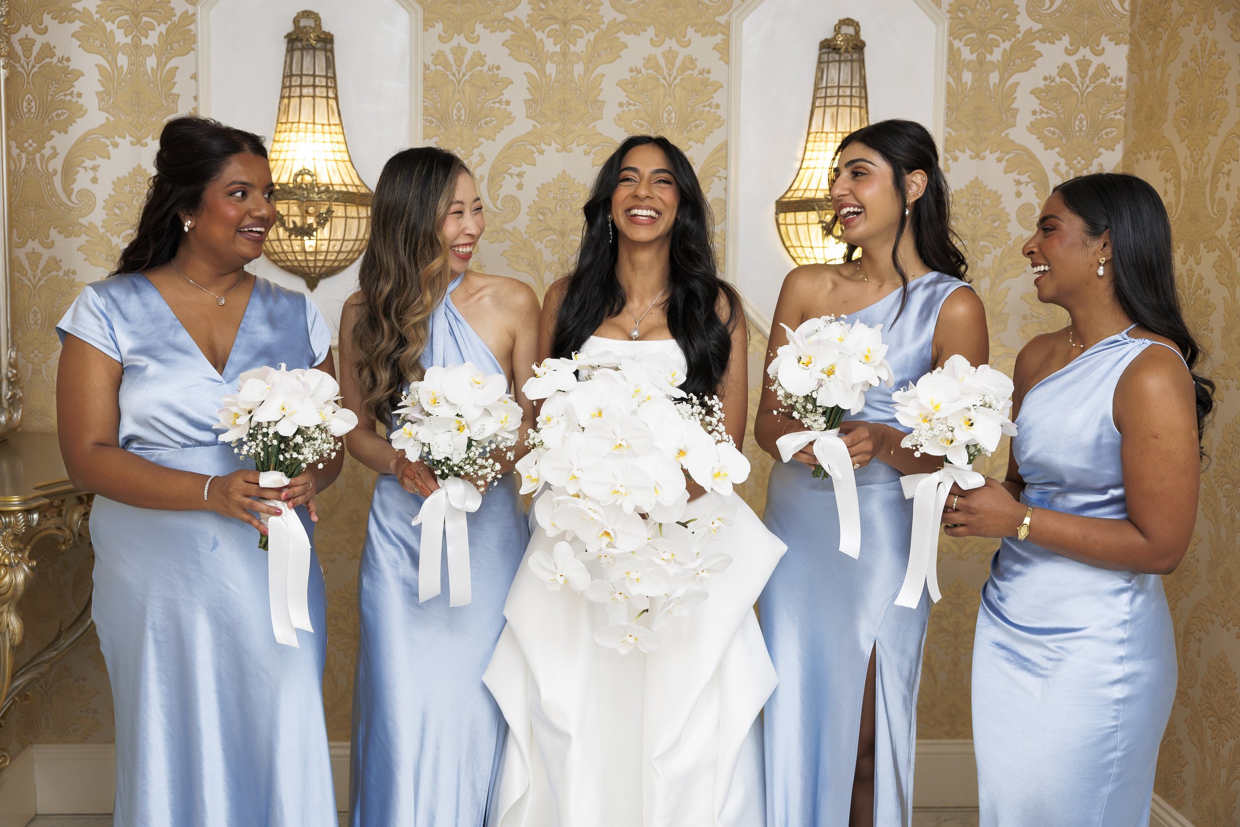 A bride with black hair and a white dress holding a cascading bouquet of white orchids, smiling in a room with gold and cream wallpaper, surrounded by five bridesmaids in matching blue dresses holding smaller bouquets, all smiling and looking at each