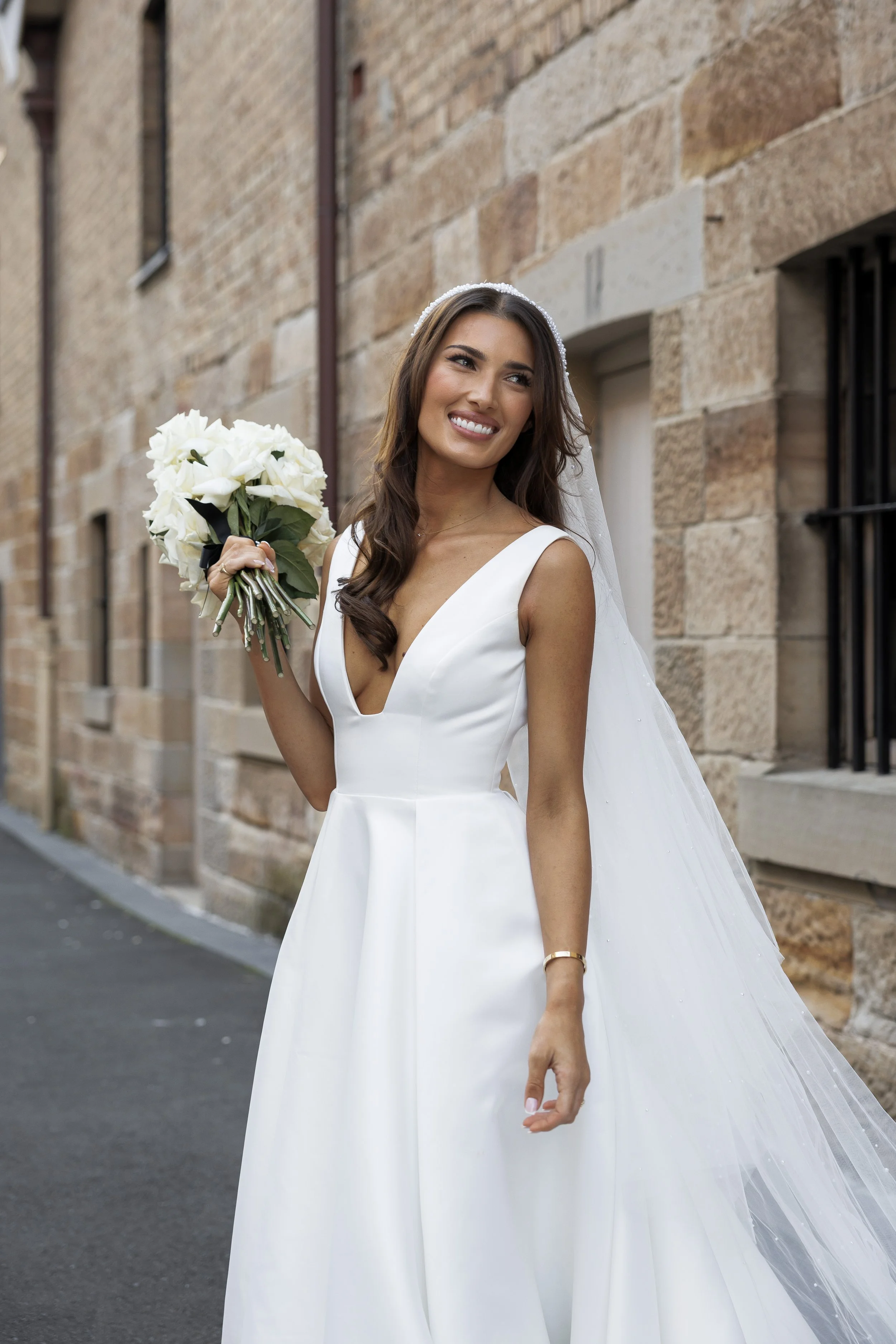 Bride in a white wedding dress holding a bouquet of white flowers, standing outside near a brick wall, smiling.