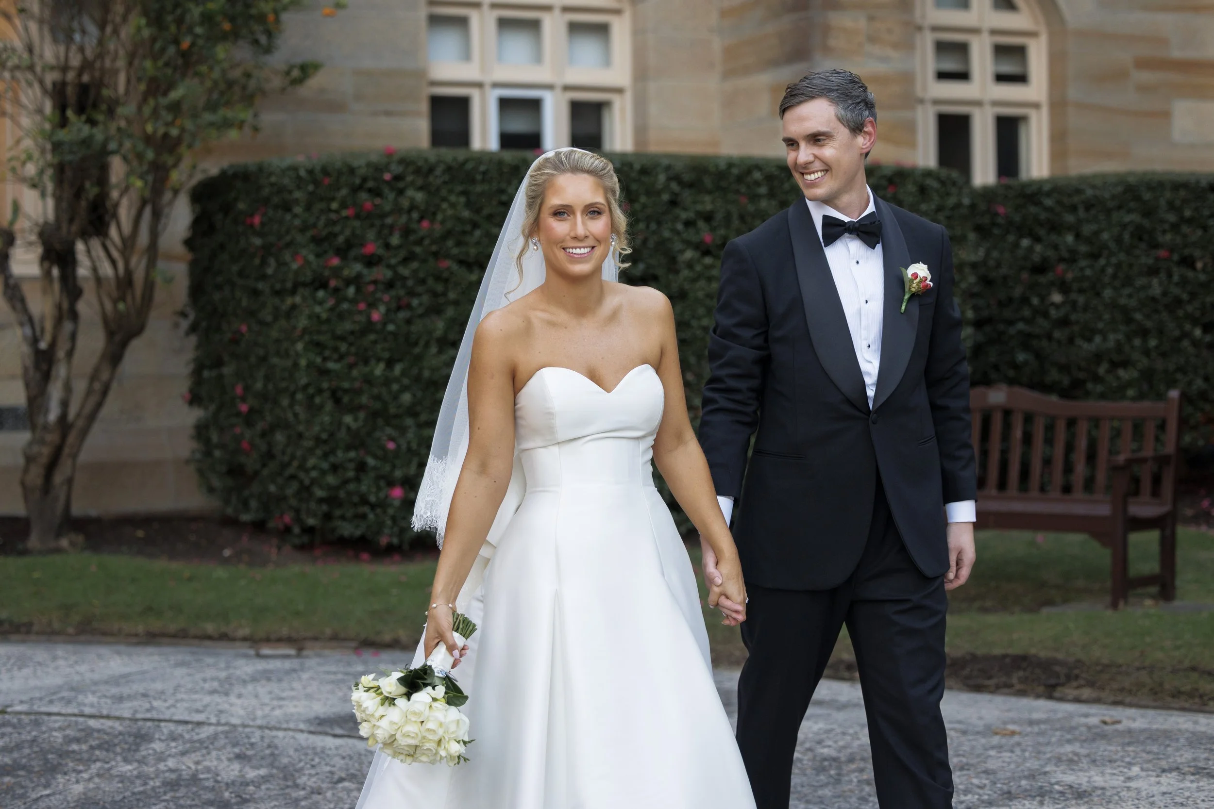 A happy bride and groom holding hands while walking outdoors, with the bride holding a bouquet of white roses, in front of a bush with pink flowers and a building in the background.