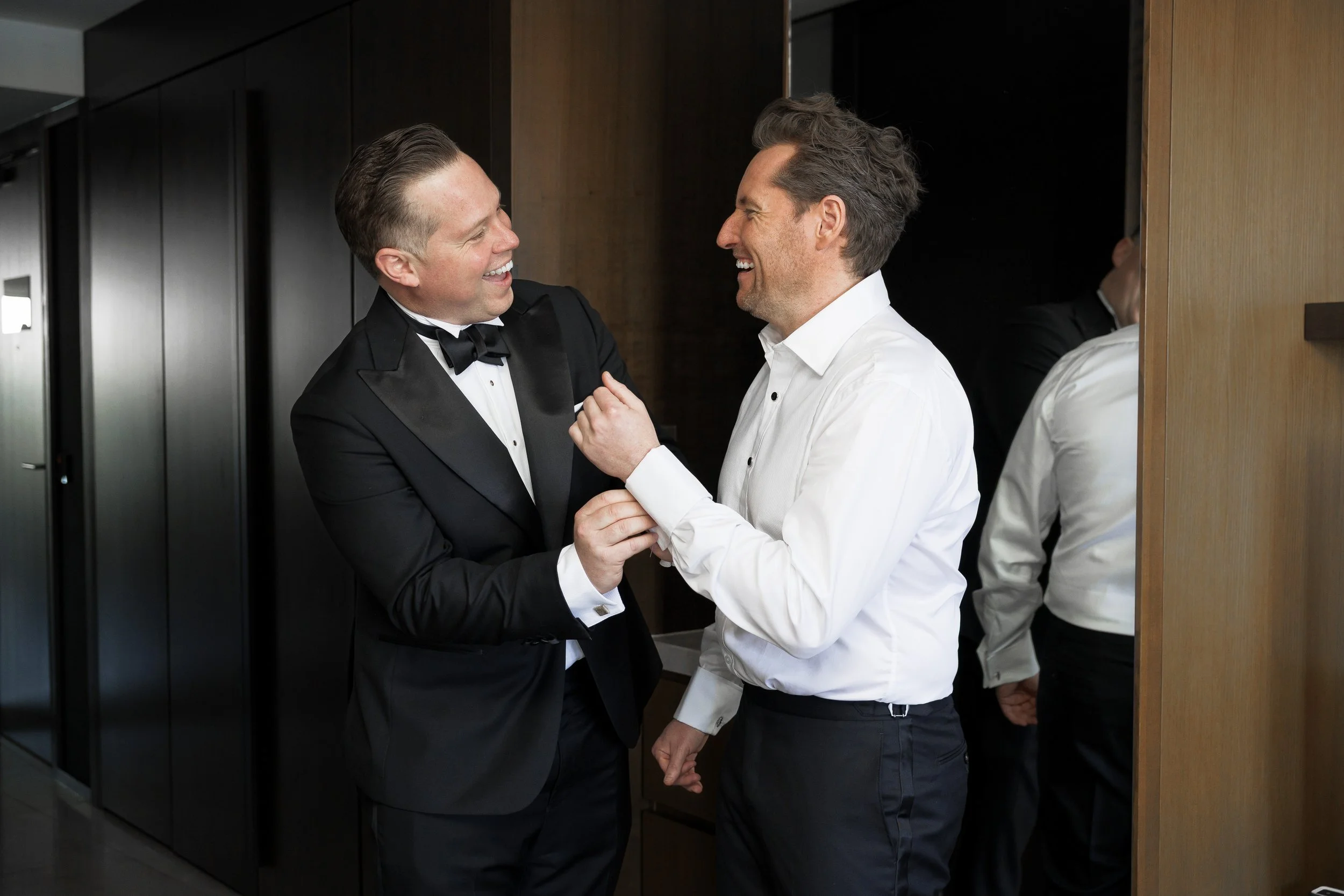 Two men laughing and enjoying each other's company, one in a tuxedo and bow tie, the other in a white dress shirt, standing in a modern indoor setting.