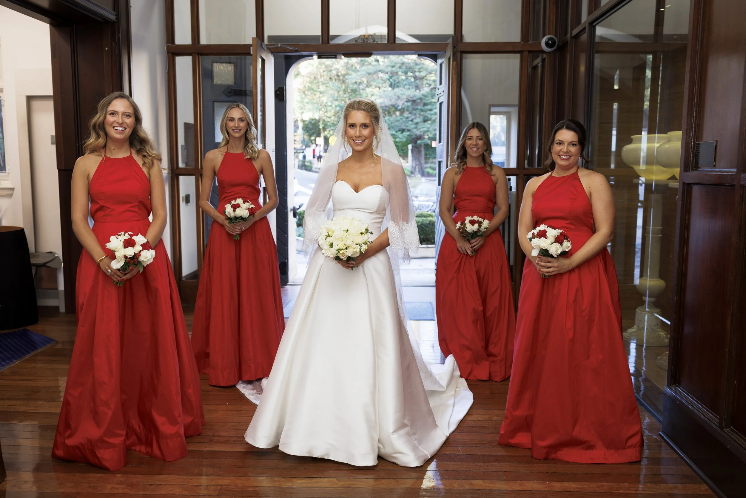A bride in a white wedding dress with a veil holding a bouquet of white flowers, standing in the center, surrounded by five bridesmaids in red dresses holding smaller bouquets, inside a room with wooden floors and a glass door background.