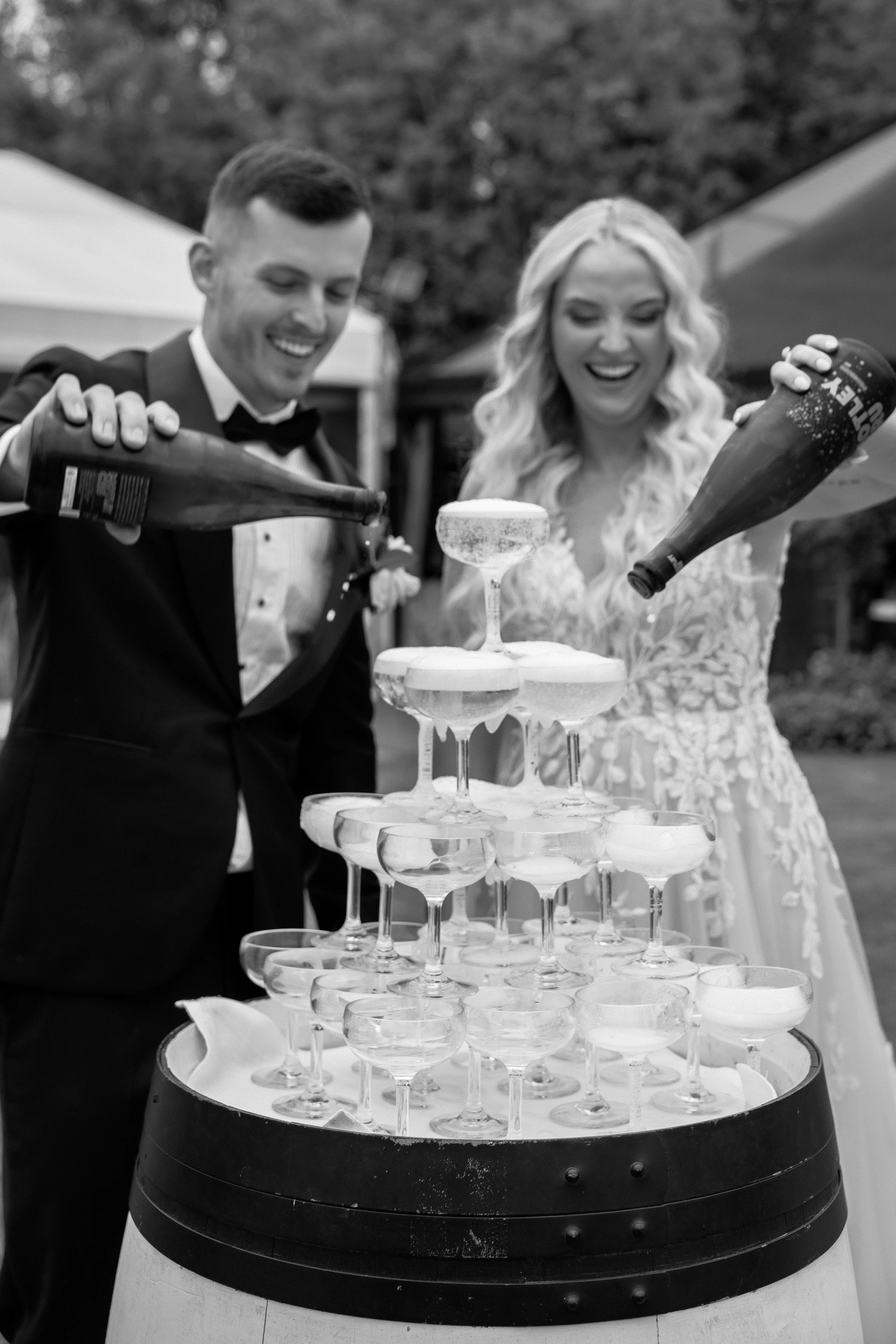A couple in formal attire pouring champagne into a tower of glass coupe glasses at an outdoor celebration.