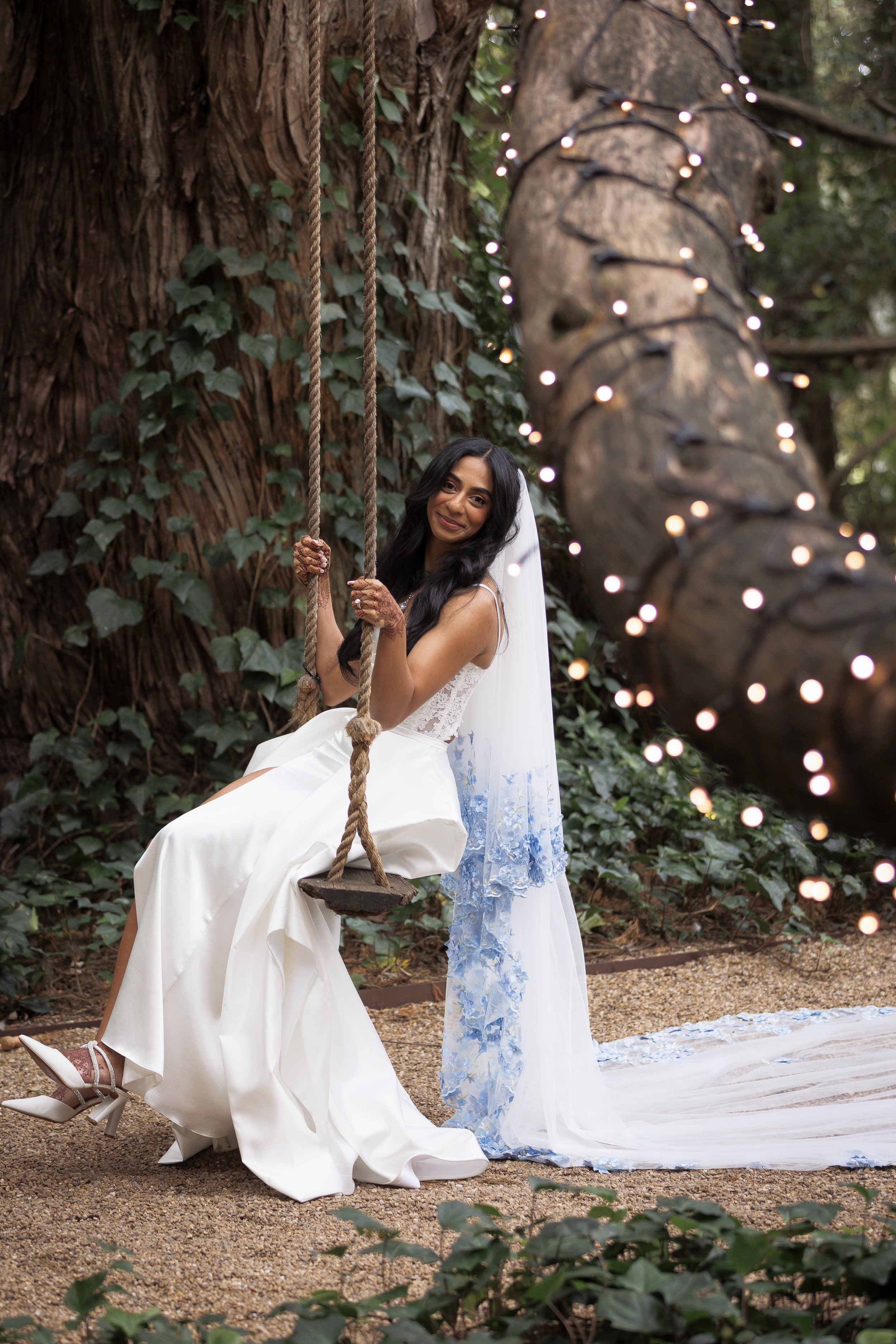 A bride in a white wedding dress sitting on a wooden swing tied to a large tree with string lights wrapped around the trunk, surrounded by lush greenery.