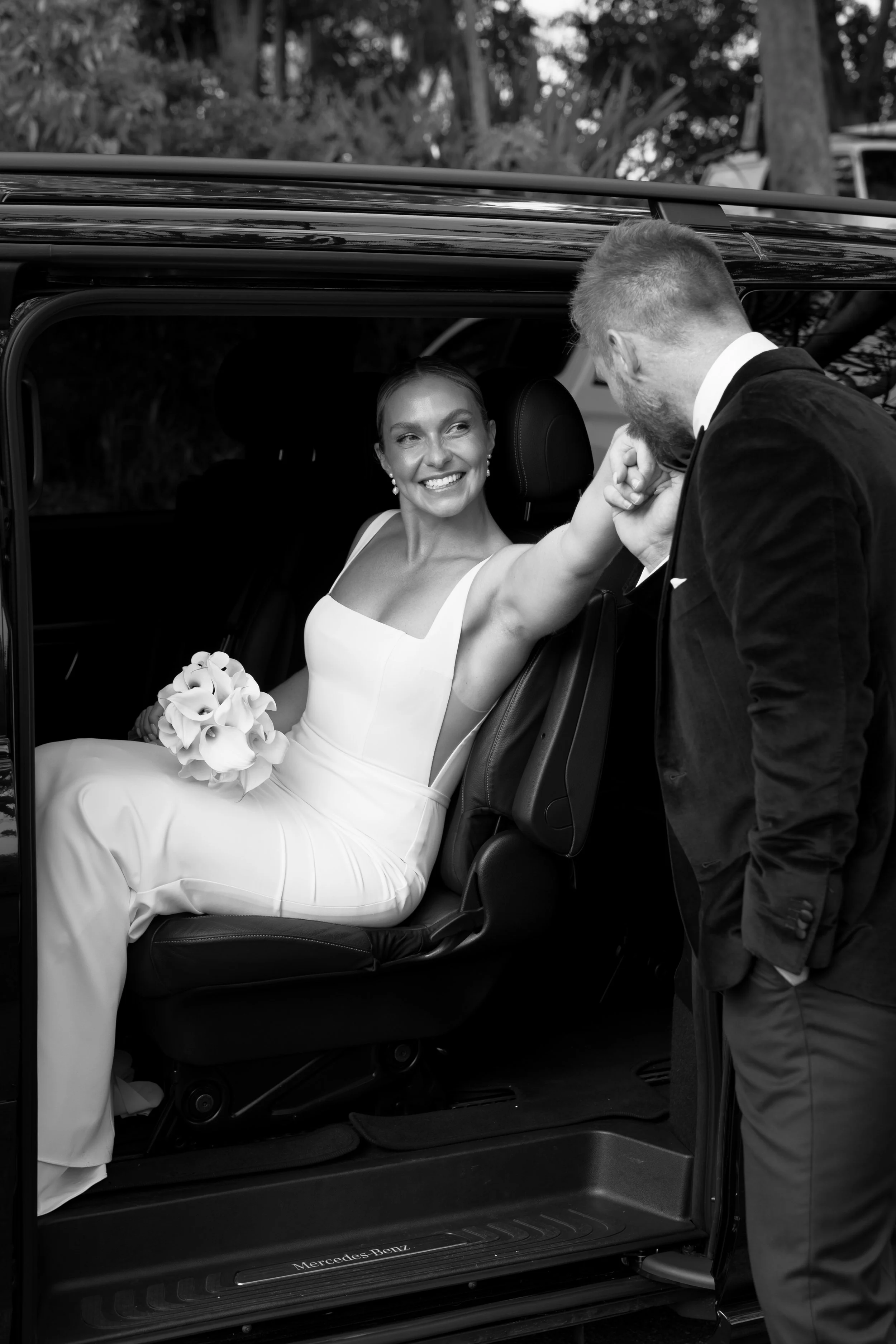 A bride in a wedding dress smiling inside a car, holding a bouquet, while a man outside the car whispers to her.