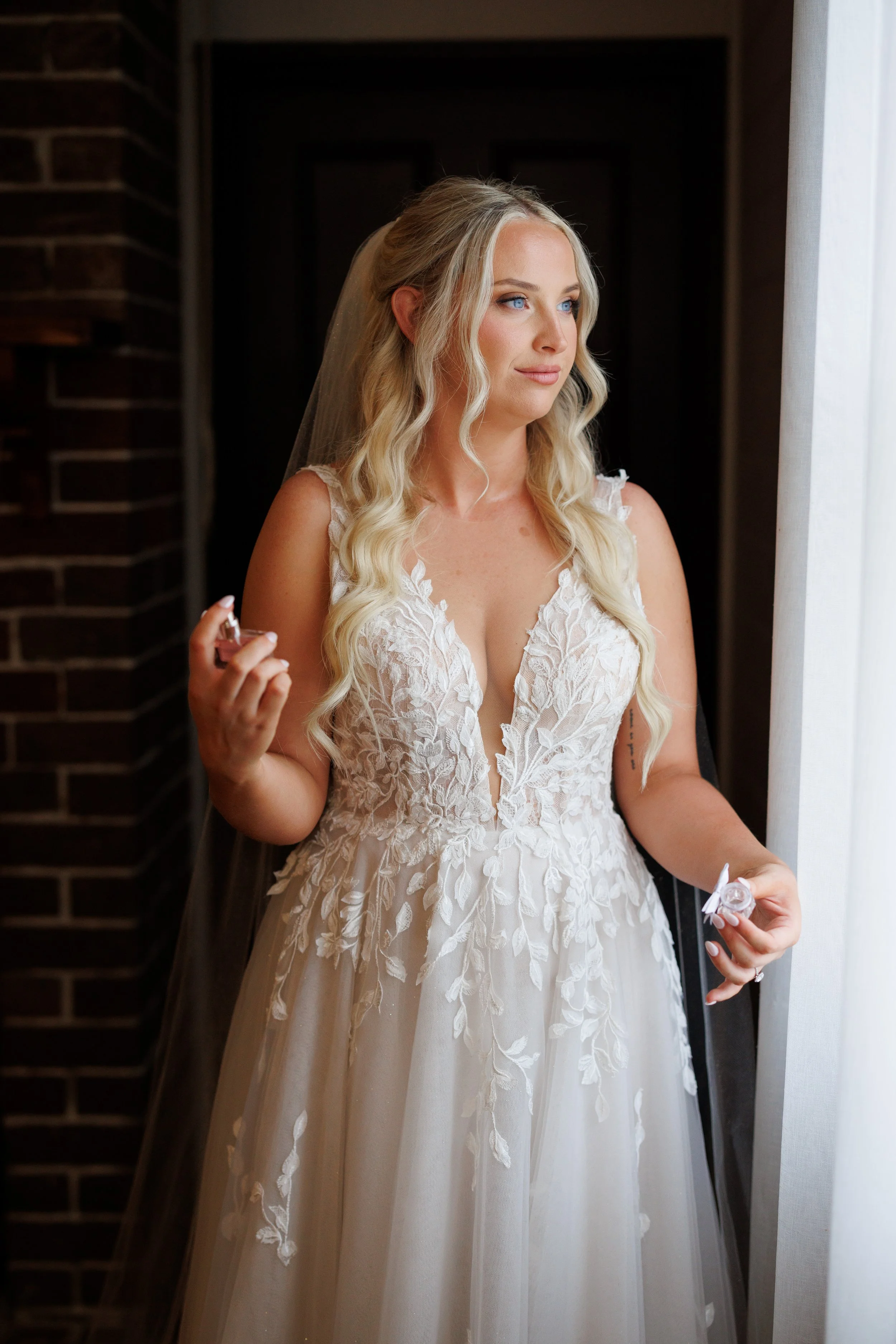 A bride with long blonde hair in loose curls, wearing a white lace wedding gown with a deep V-neck and floral embroidery, standing by a window with sheer white curtains, holding wedding rings in her hands and looking thoughtfully outside.