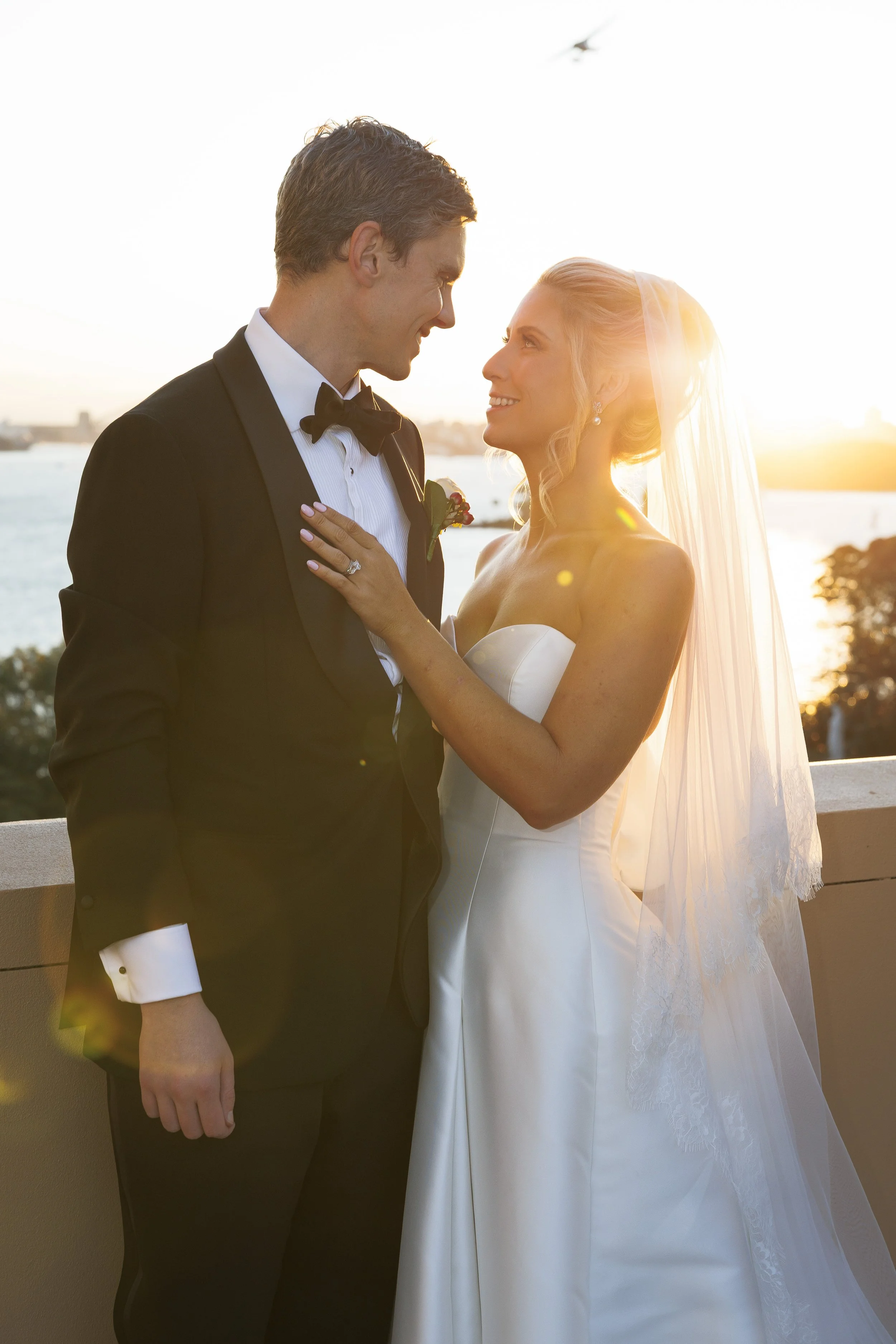 A bride and groom in wedding attire sharing a tender moment at sunset with water and city skyline in the background.