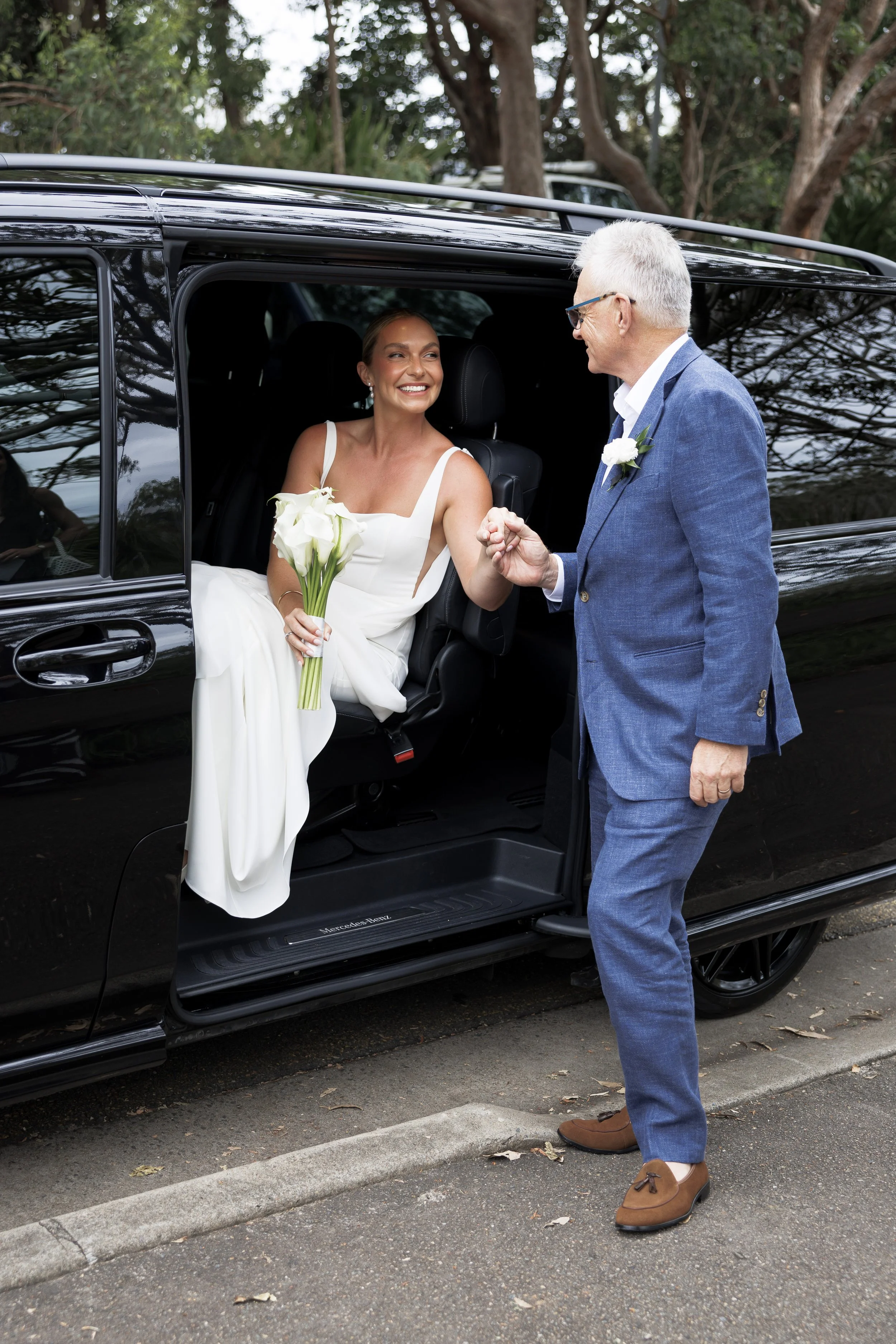 A bride in a white wedding dress holding a bouquet of white flowers, sitting in a black vehicle, smiling and holding hands with an older man in a blue suit, standing outside next to the vehicle, holding her hand.