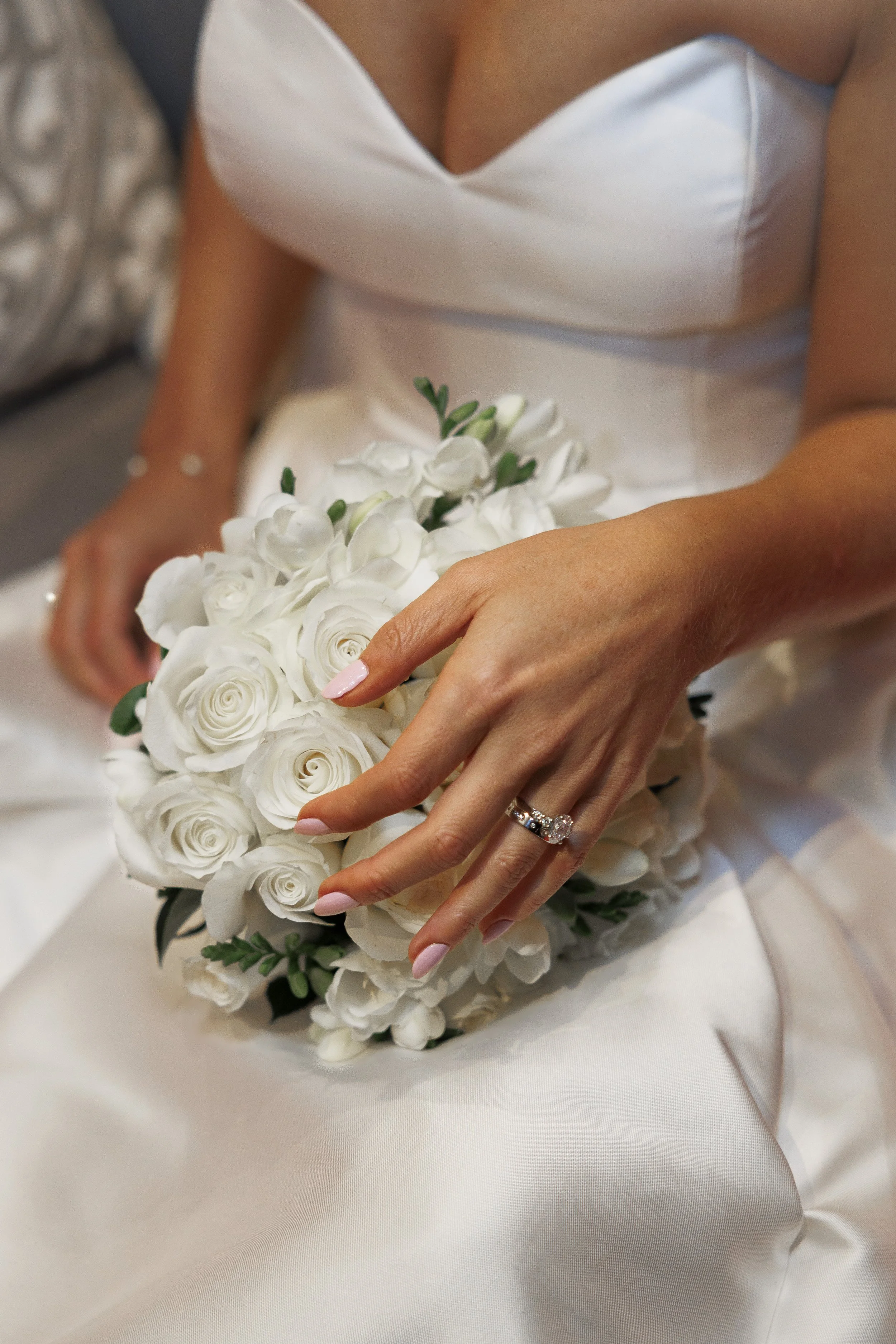 Close-up of a bride's hand with a diamond ring, holding a bouquet of white roses, while sitting in a wedding dress.