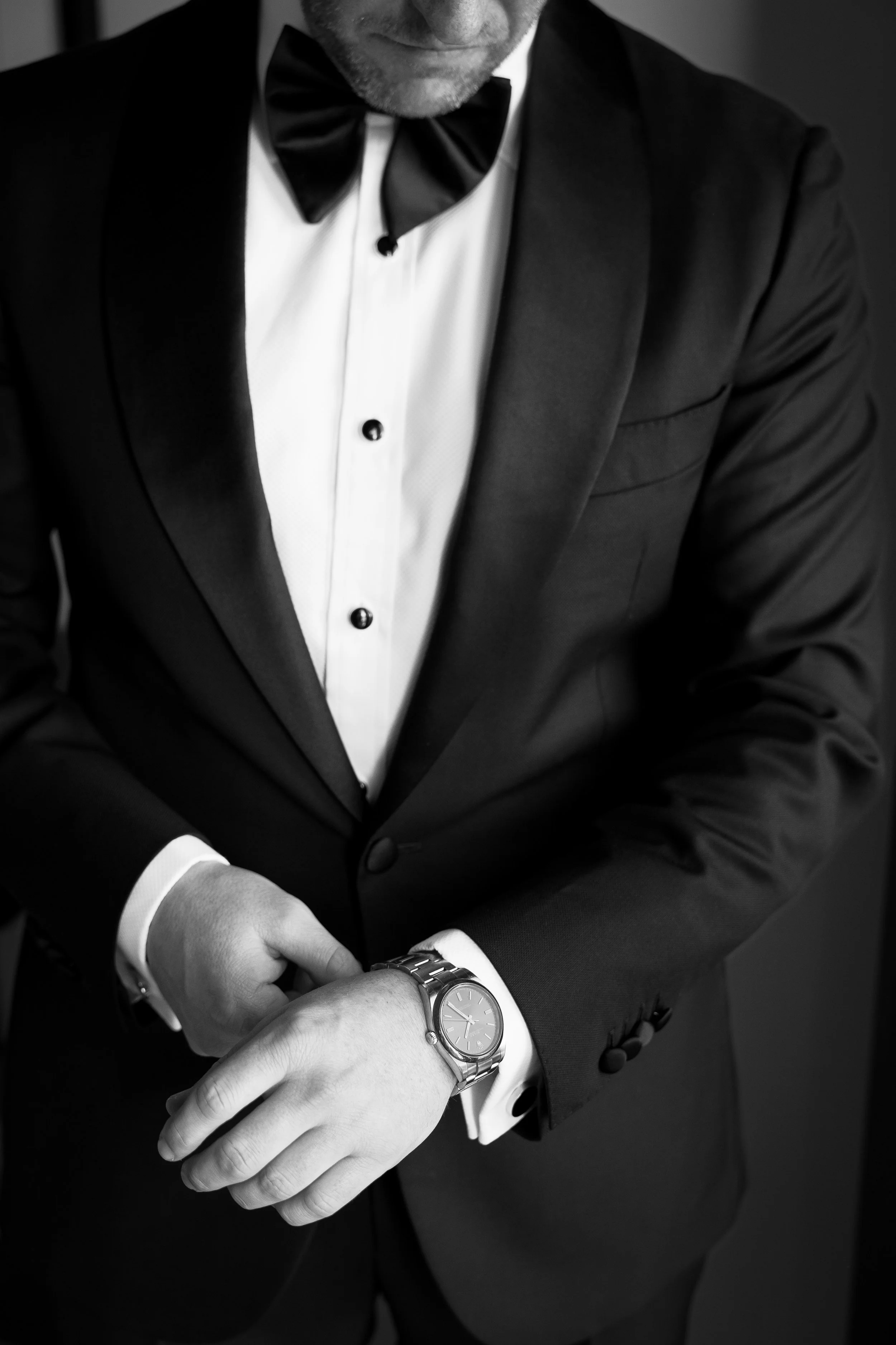 A man in a tuxedo, adjusting a wristwatch, with a black bow tie and white shirt visible, in black and white.