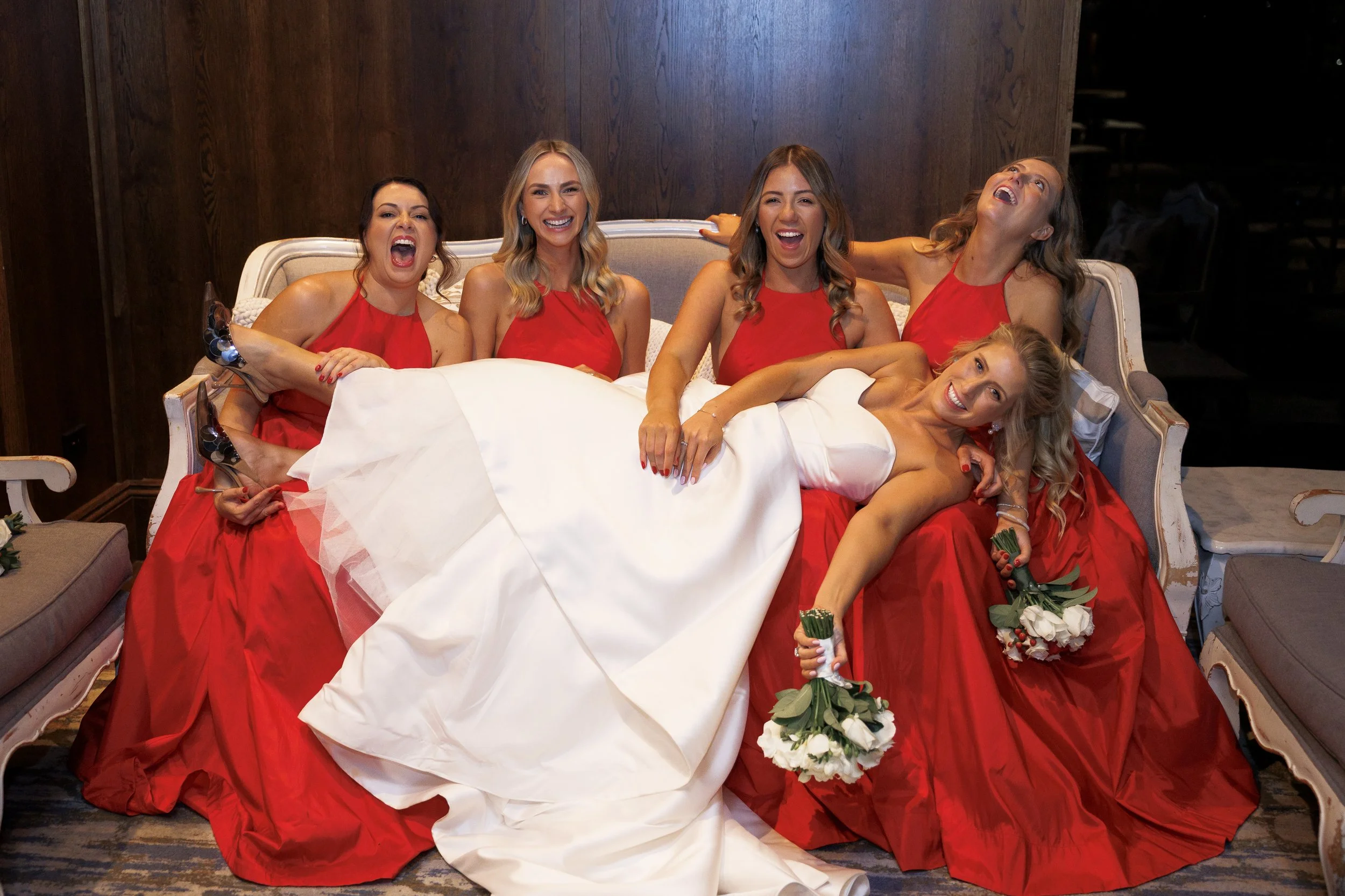 A bride in a white wedding gown is lying on a vintage-style sofa, surrounded by five bridesmaids in matching red dresses, all smiling and laughing.