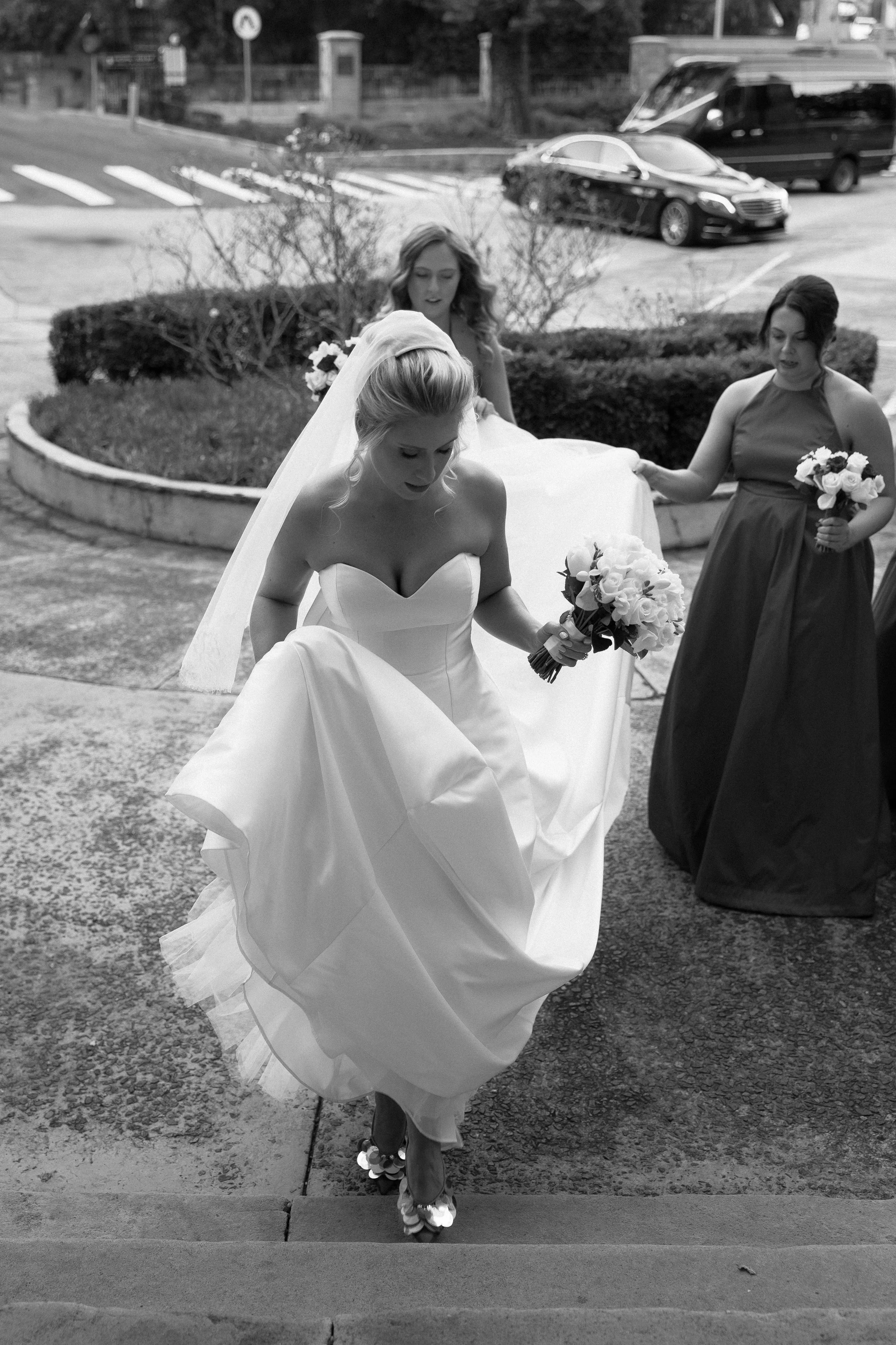 A bride in a wedding dress and veil holding a bouquet, walking up stairs with bridesmaids around her, some holding bouquets, outside a building.