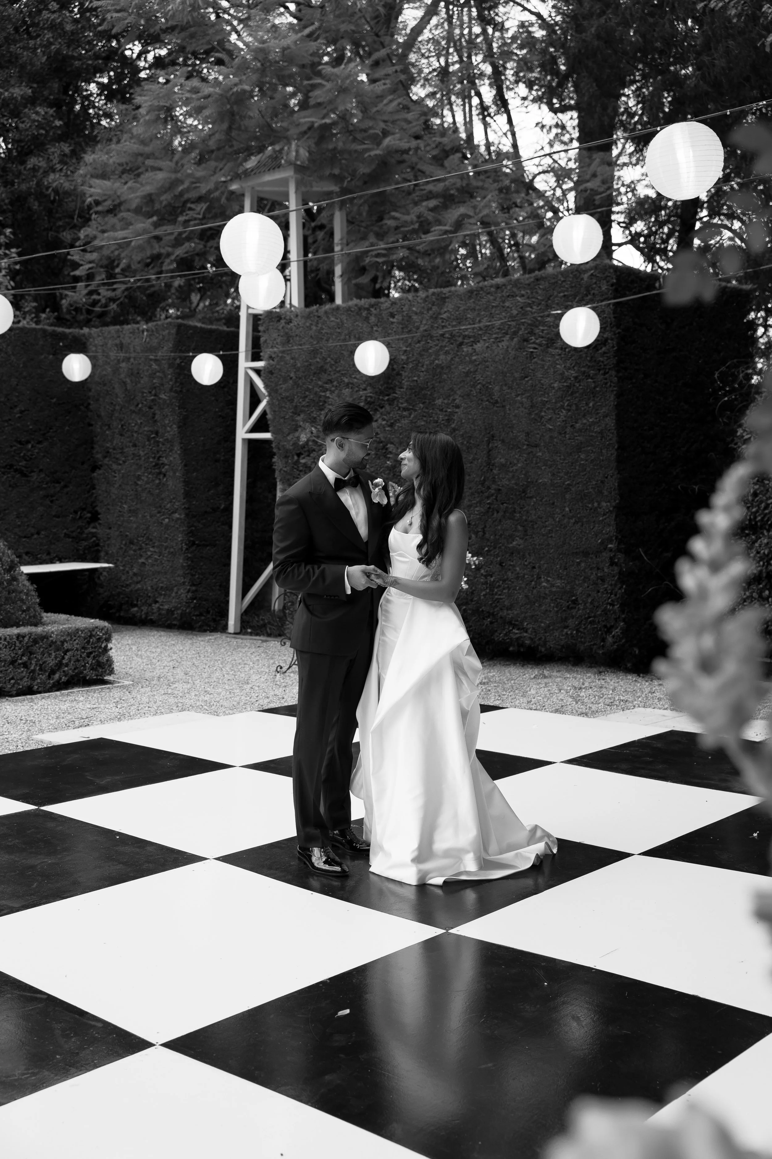 Black and white photo of a bride and groom dancing on a checkered dance floor outside, surrounded by hanging lanterns and tall hedges.