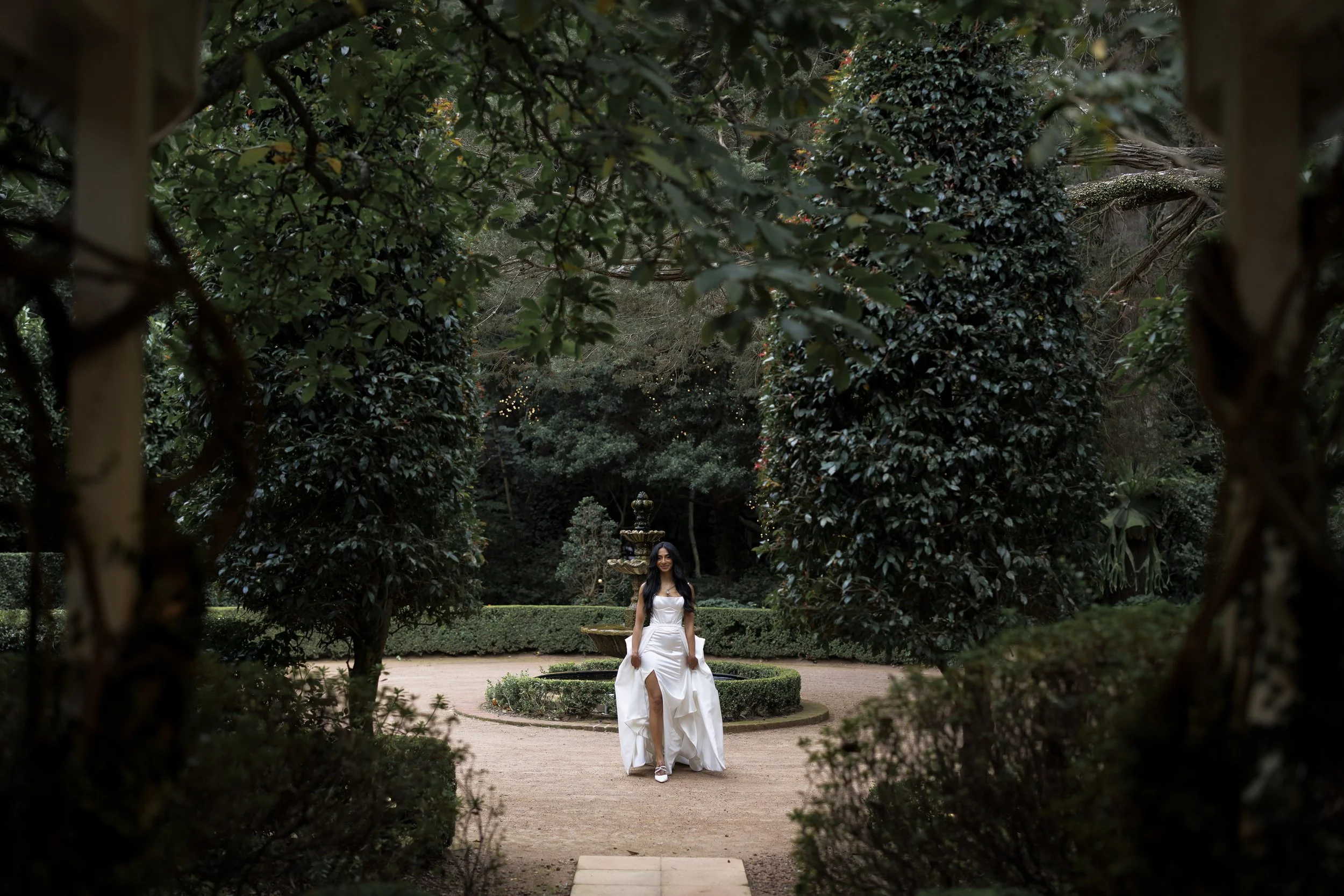 A woman in a white gown walking in a lush green garden with a fountain in the background