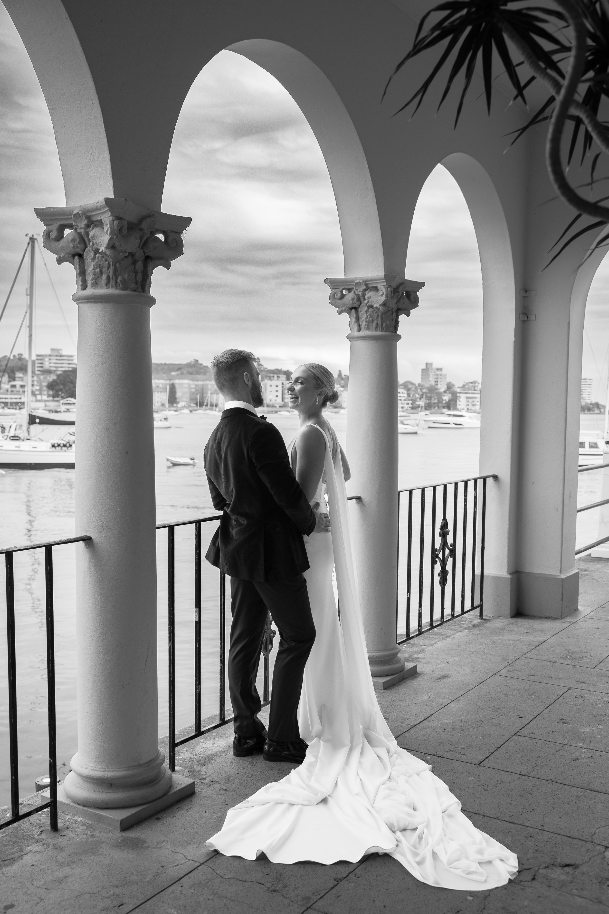 A black and white photograph of a bride and groom standing on a balcony with arches, overlooking a marina with boats and a city skyline in the background. The couple is smiling and holding each other, with the bride wearing a long, flowing gown and t