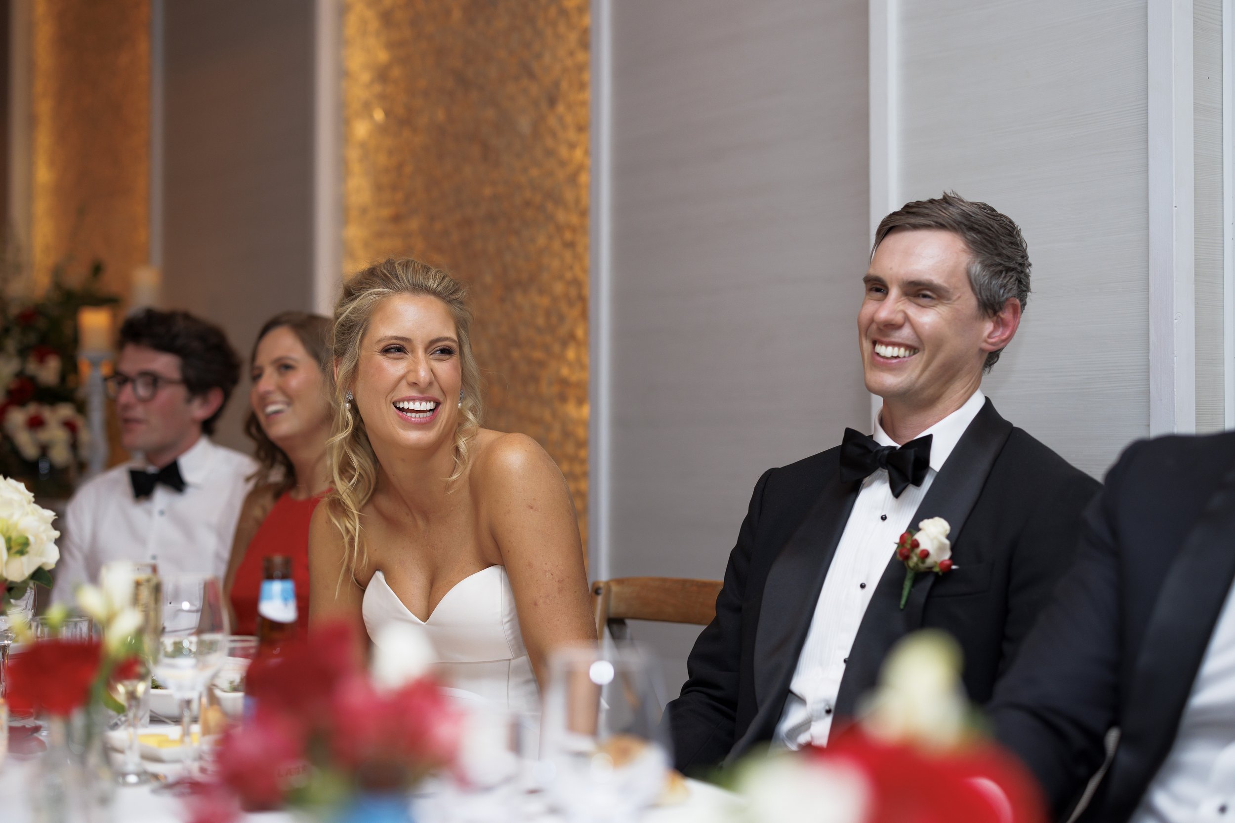 People at a wedding reception, including a woman in a white dress and a man in a tuxedo, smiling and laughing.