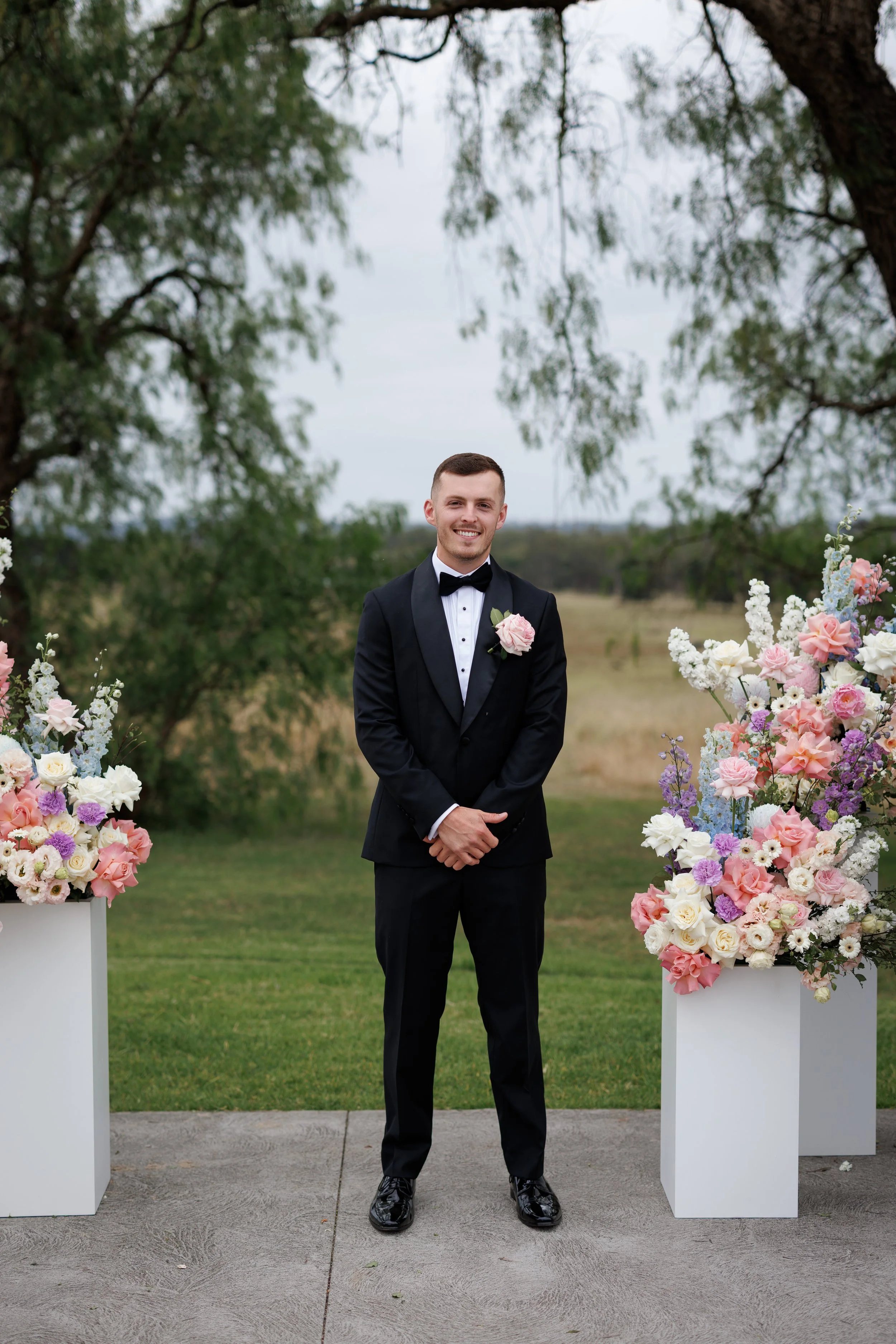 A groom in a black tuxedo with a pink boutonniere stands outdoors between two large floral arrangements, smiling at the camera.