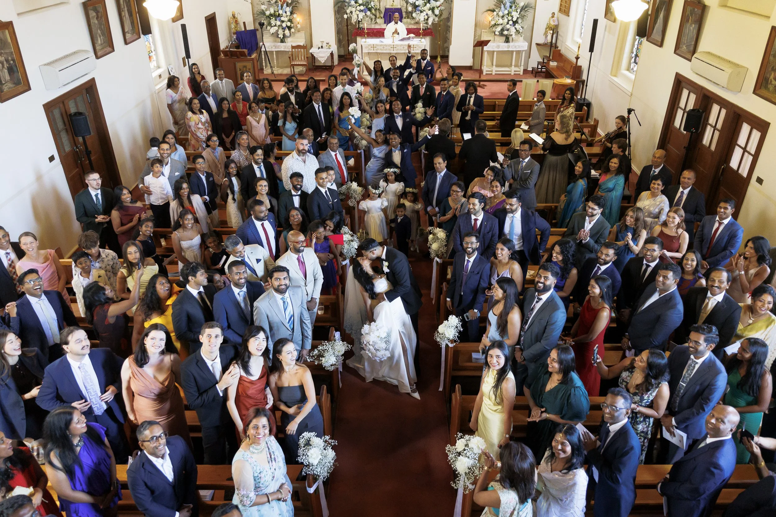 A joyful wedding ceremony inside a church with a bride and groom kissing at the altar, surrounded by numerous guests seated in pews, some standing and smiling, and floral decorations.