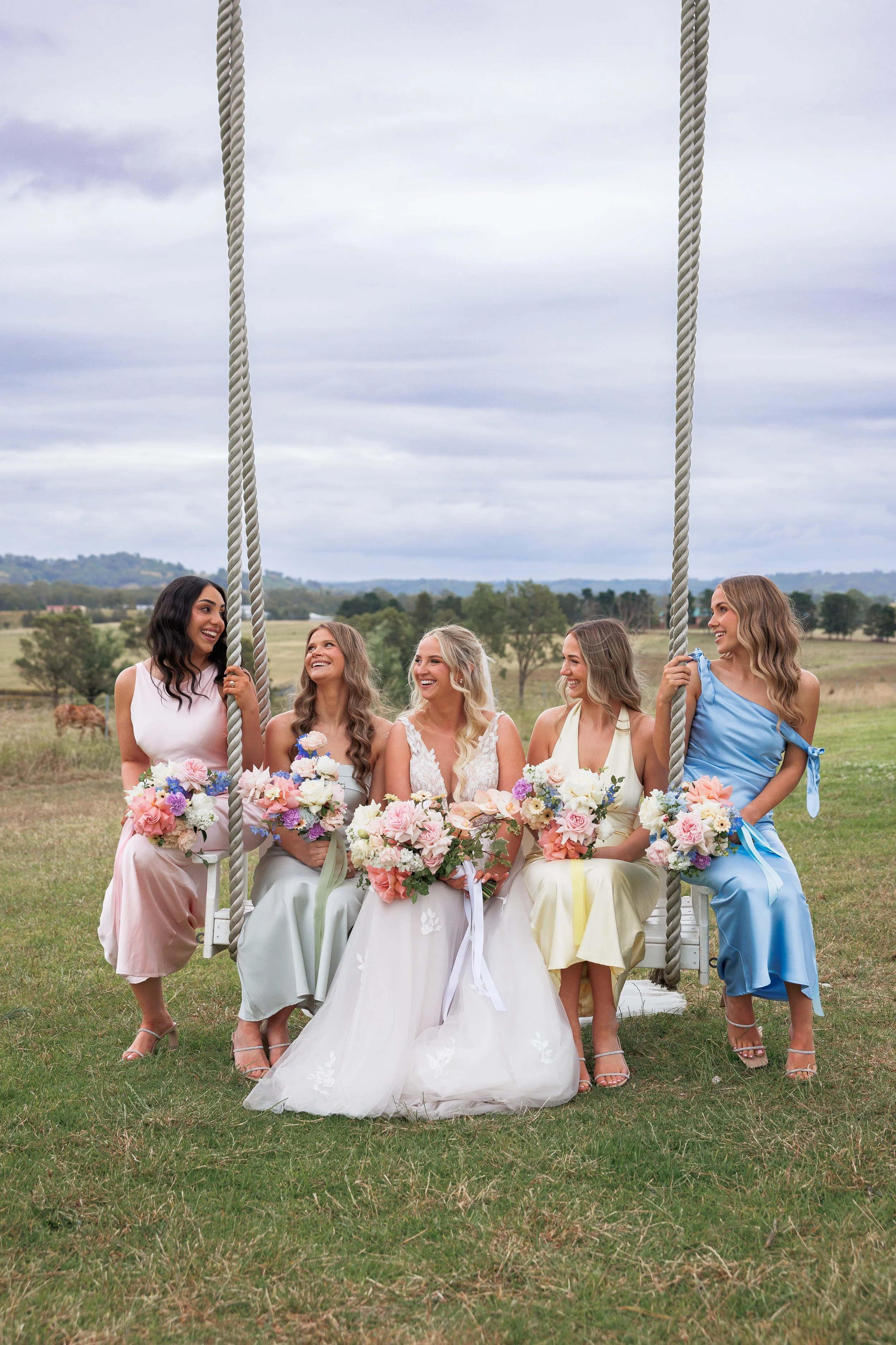 Five women in colorful dresses sitting on a white swing outdoors, holding bouquets, smiling, with a rural landscape and cloudy sky in the background.