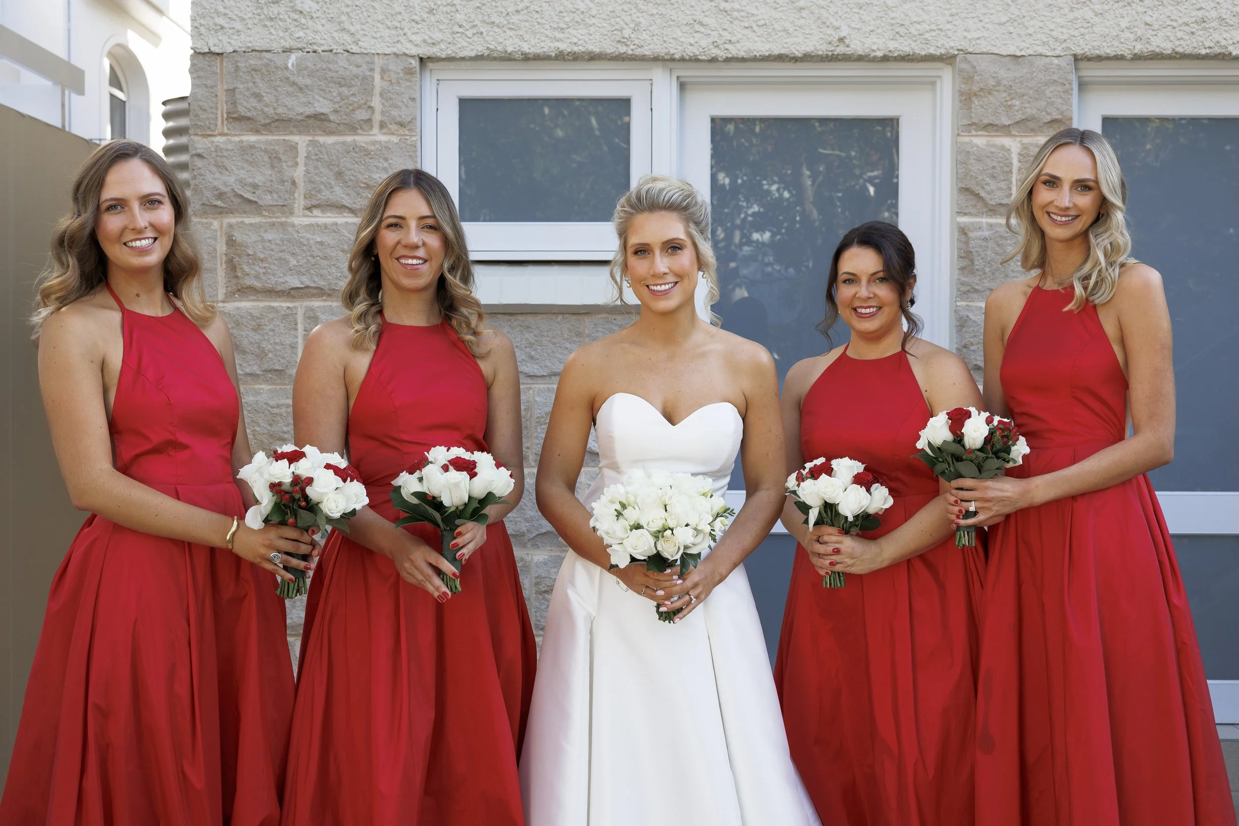 Bride in a white strapless wedding dress holding a bouquet of white roses, standing with four bridesmaids in red dresses, each holding a bouquet of red and white roses.