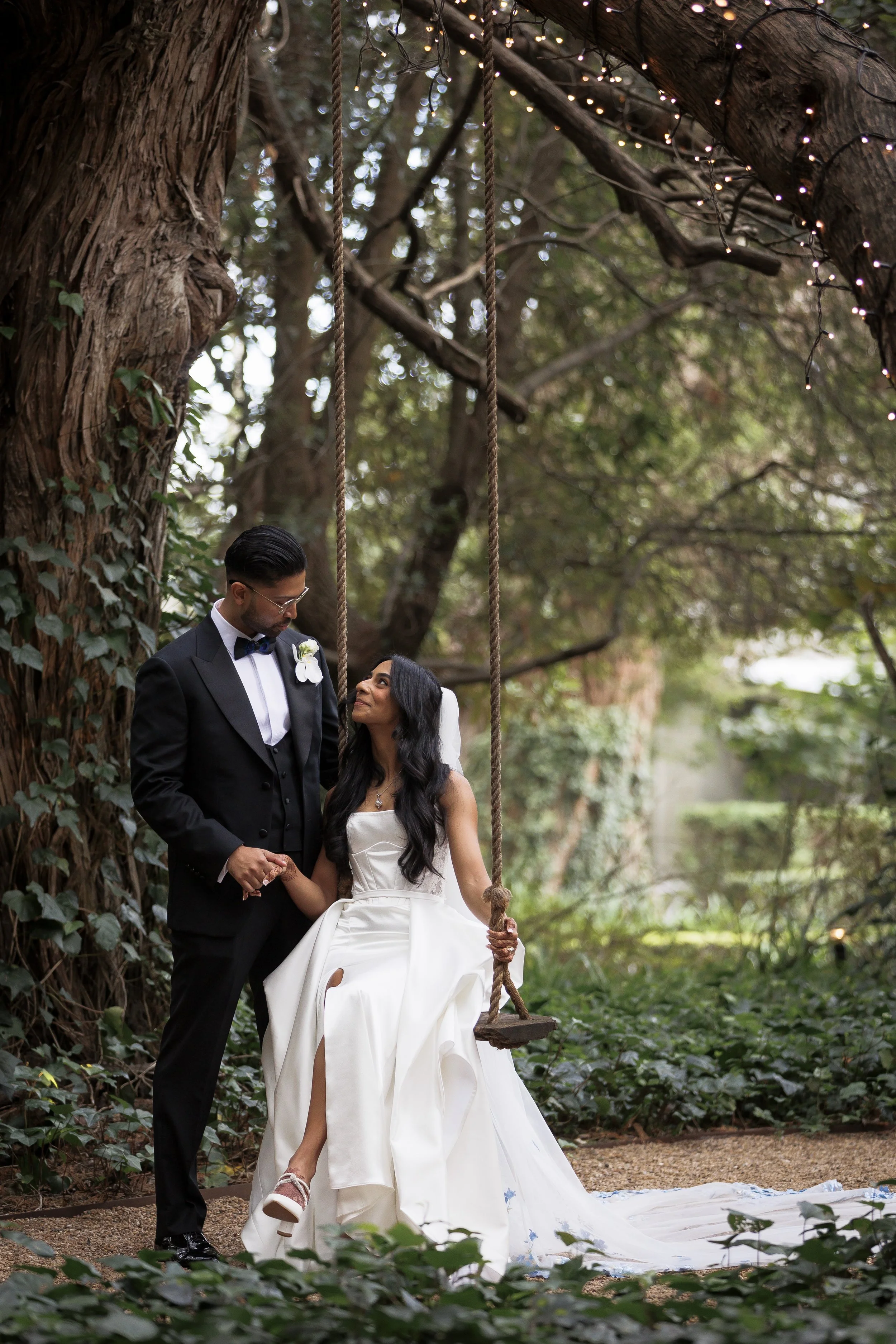 A bride and groom in wedding attire standing together outdoors near a large tree with a swing hanging from its branches, surrounded by greenery and string lights.