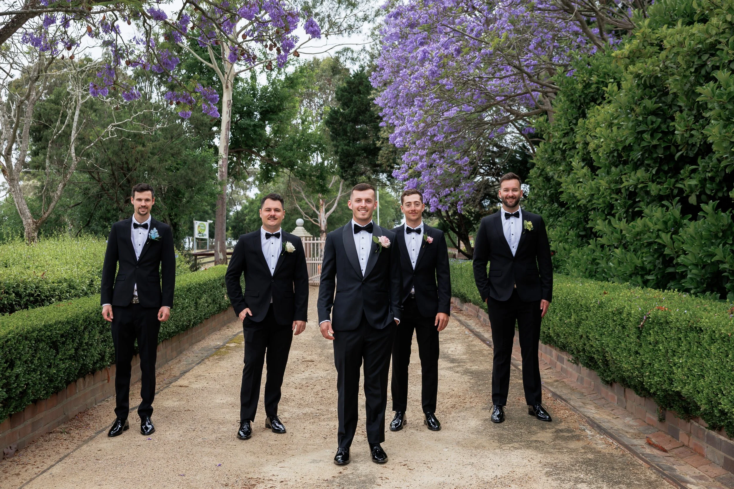 Five men in tuxedos walking down a garden path, with purple flowering trees and green bushes in the background.